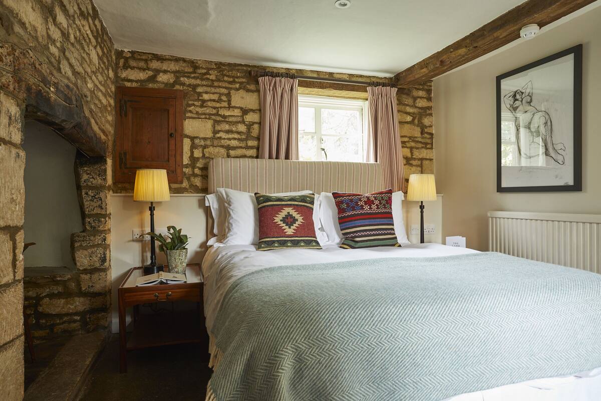 A bedroom with a stone wall, a bed with patterned pillows, a nightstand with a lamp, and a framed artwork on the wall.
