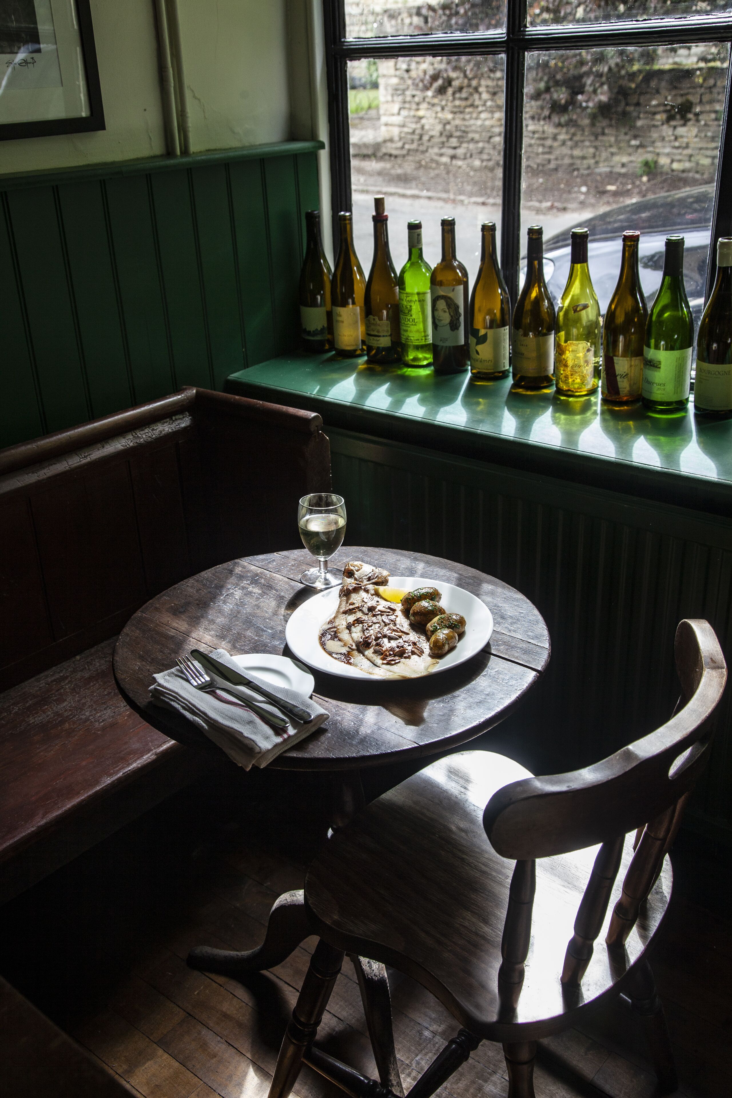 A dining table with a plate of food and a glass of wine, set against a window with bottles of wine on the sill.
