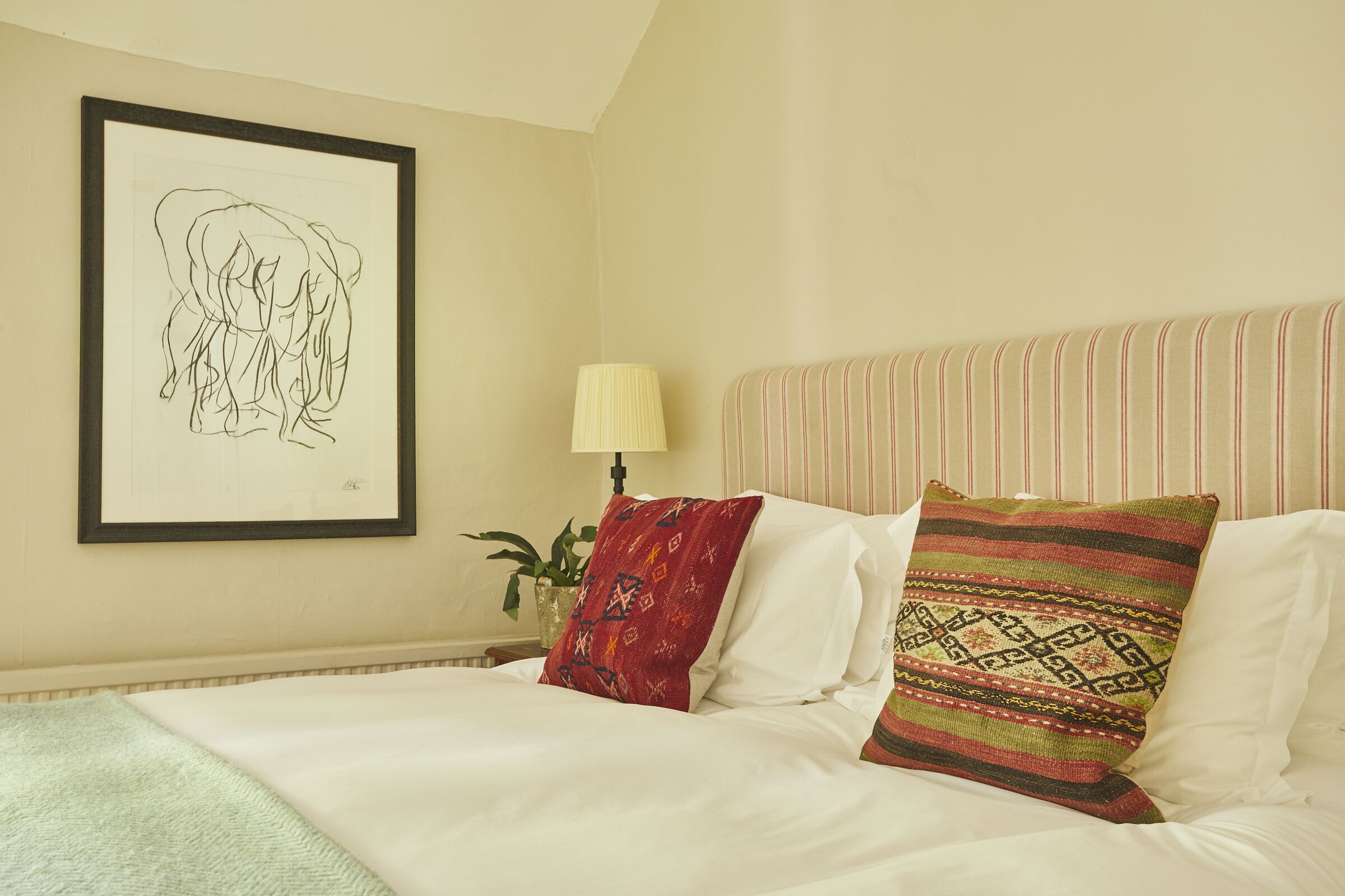 Een net opgemaakt bed met witte lakens en twee decoratieve kussens, een ingelijste afbeelding aan de muur en een bedlampje.