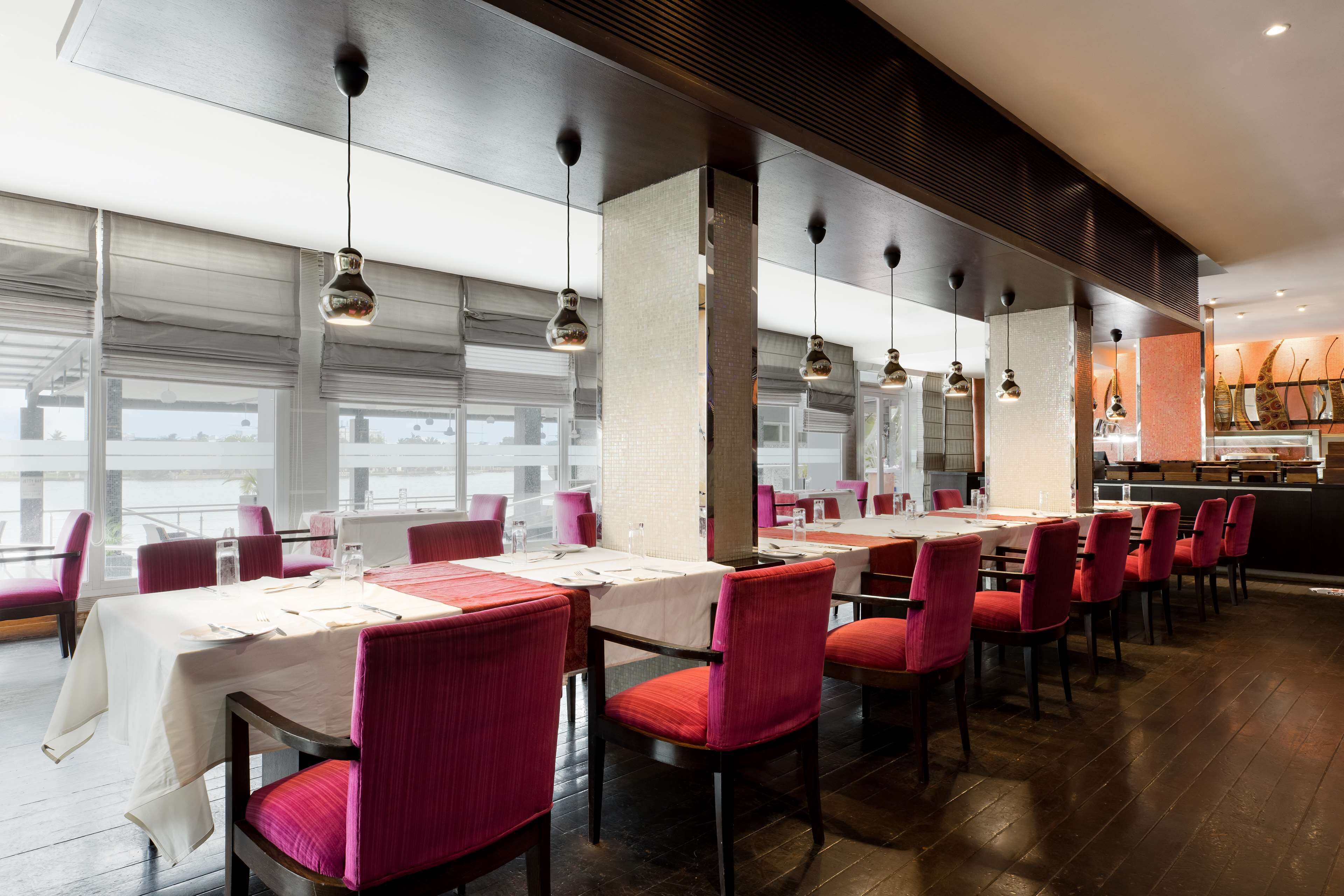 A restaurant with long tables, red chairs, and hanging pendant lights.