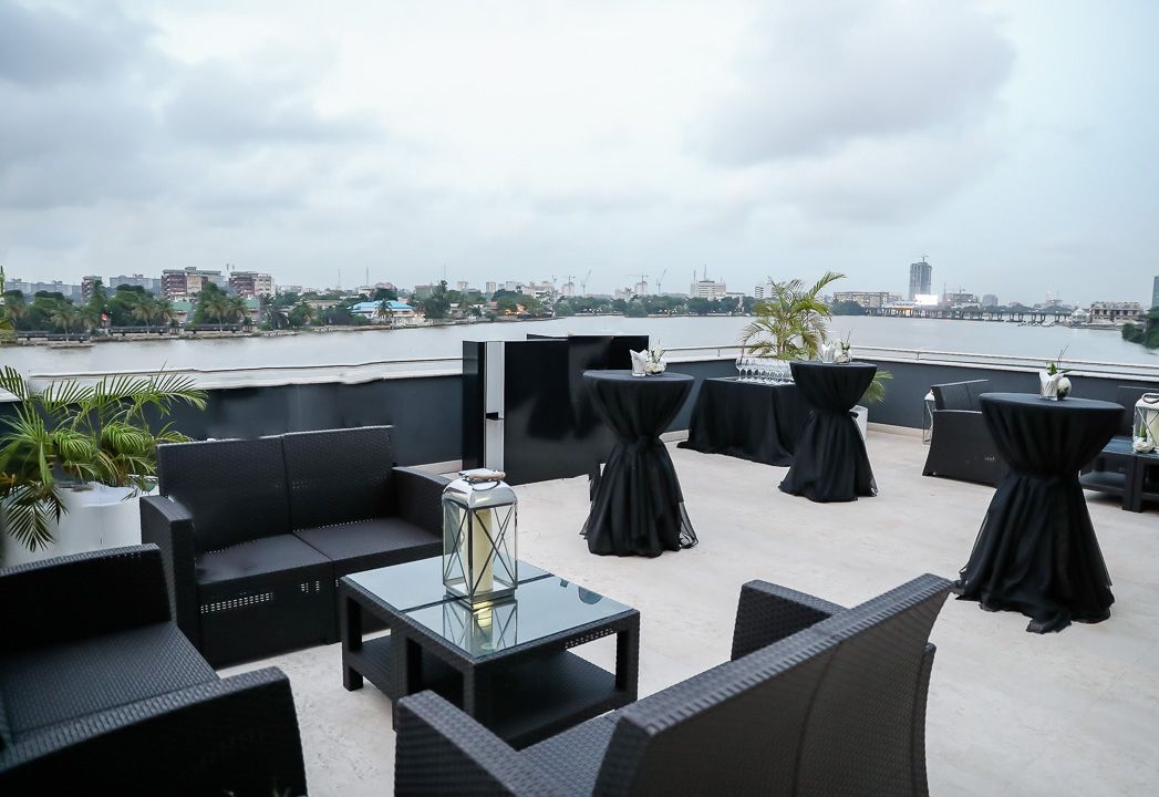 A rooftop terrace with black furniture, round tables, and potted plants.