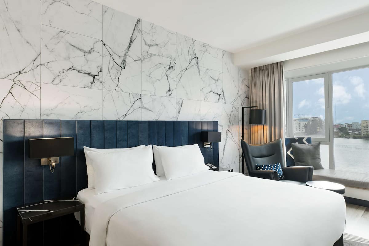 A modern hotel room with a large bed, marble-patterned wall, a view of the city, and a seating area by the window.