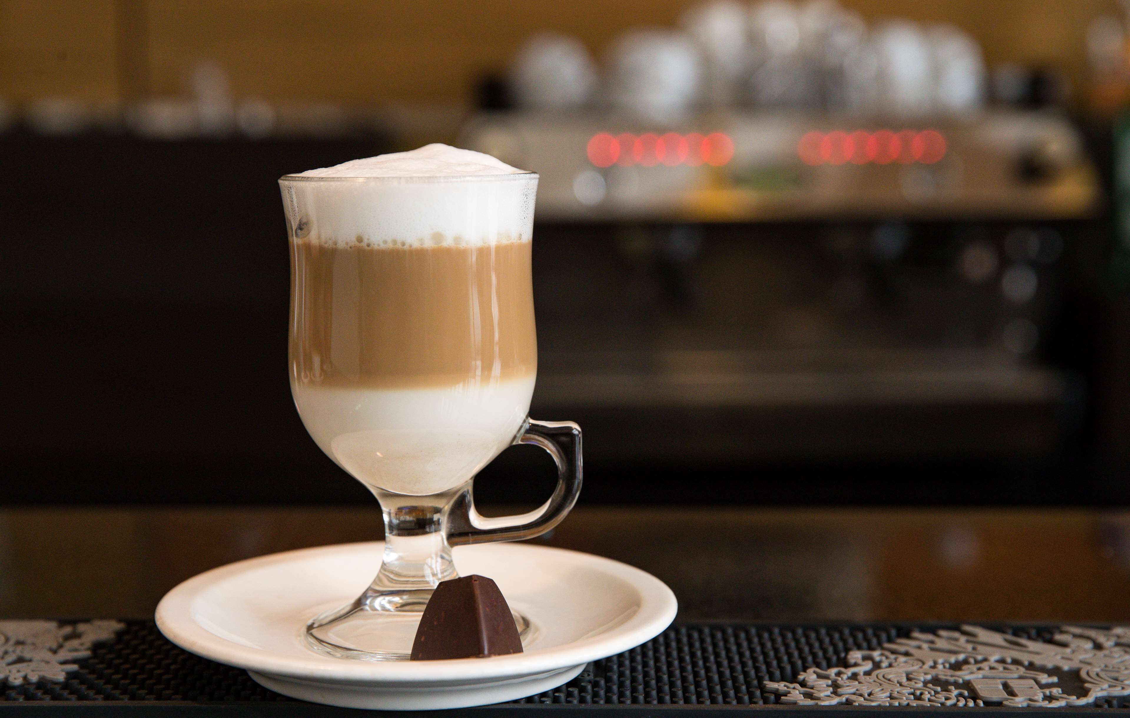 A glass of layered coffee on a saucer with a chocolate piece beside it.