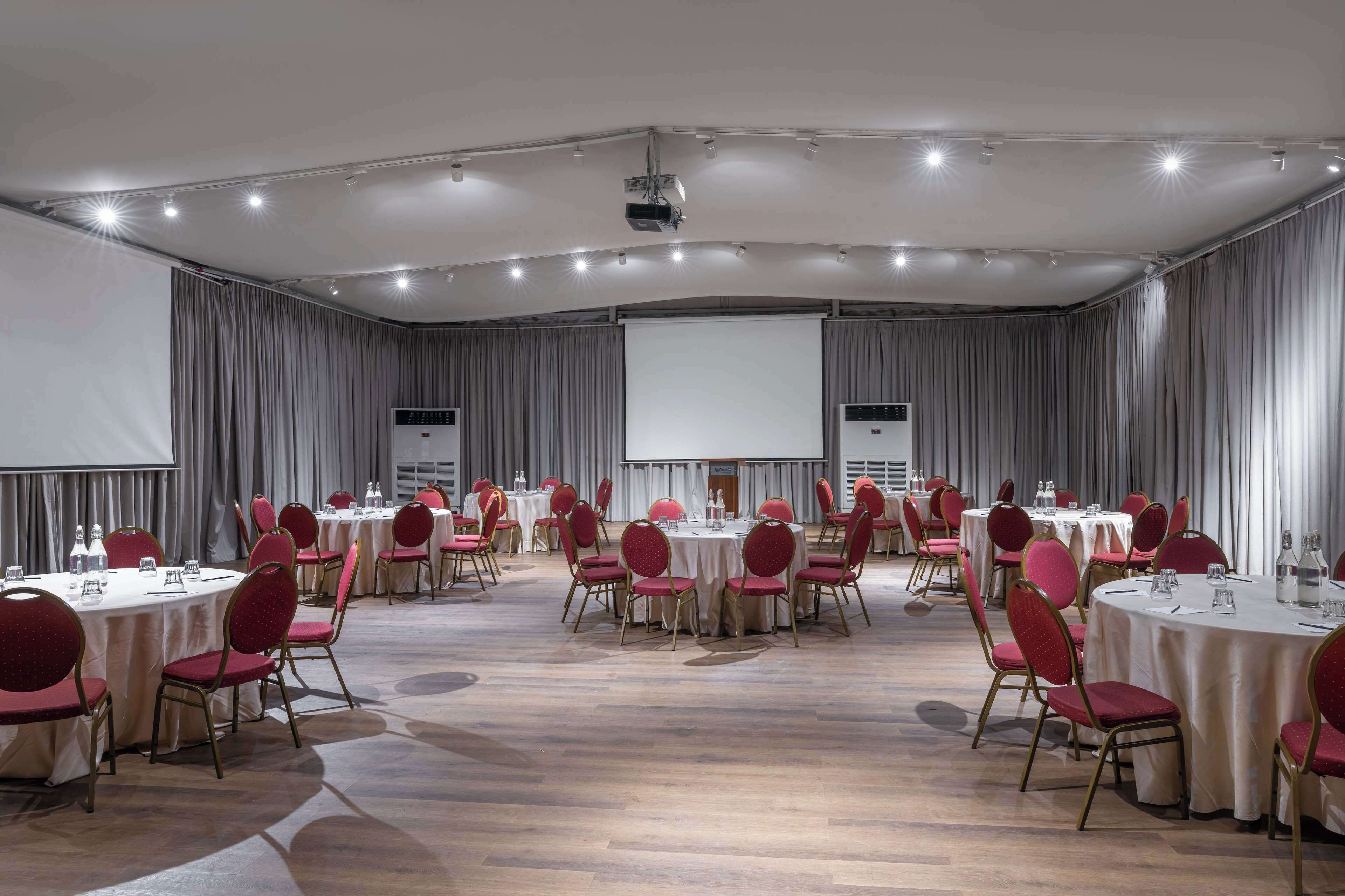 A large conference room with round tables and red chairs, equipped with projectors and screens.
