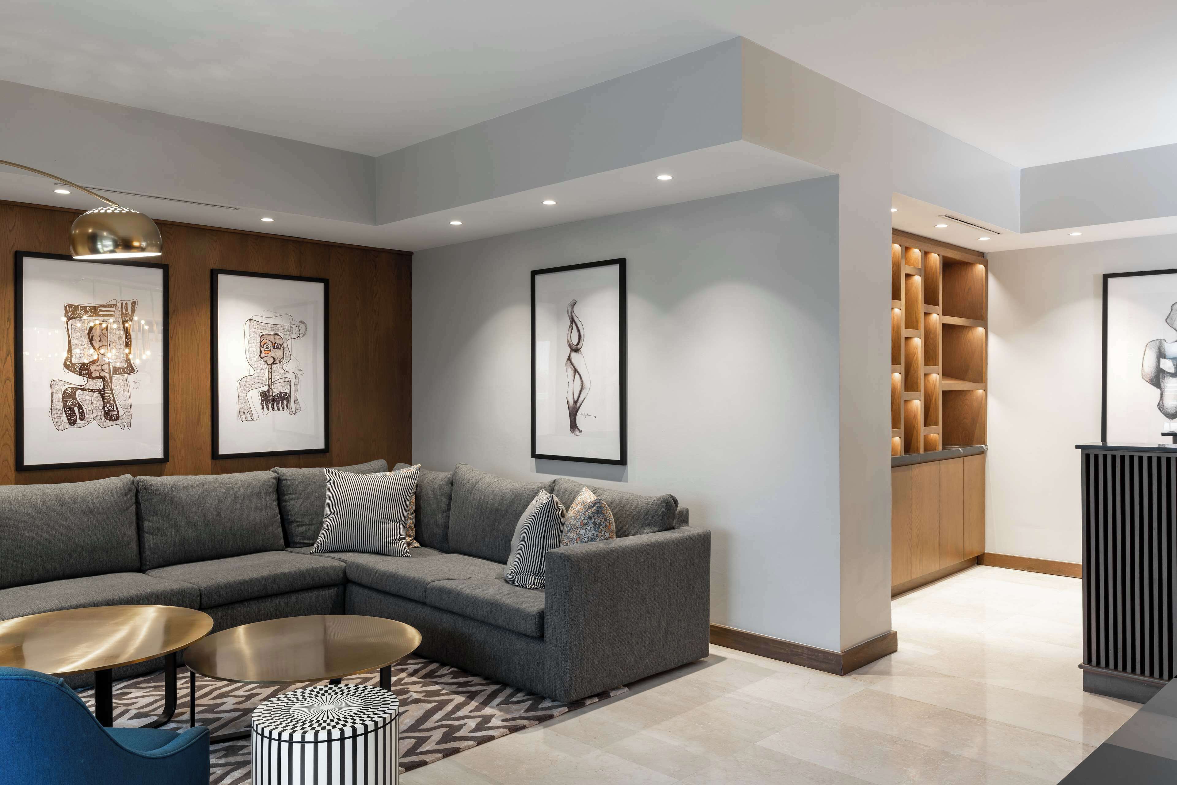A modern living room with a grey sectional sofa, a round coffee table, and a patterned rug. There are framed artworks on the walls and a built-in shelving unit with decorative items.