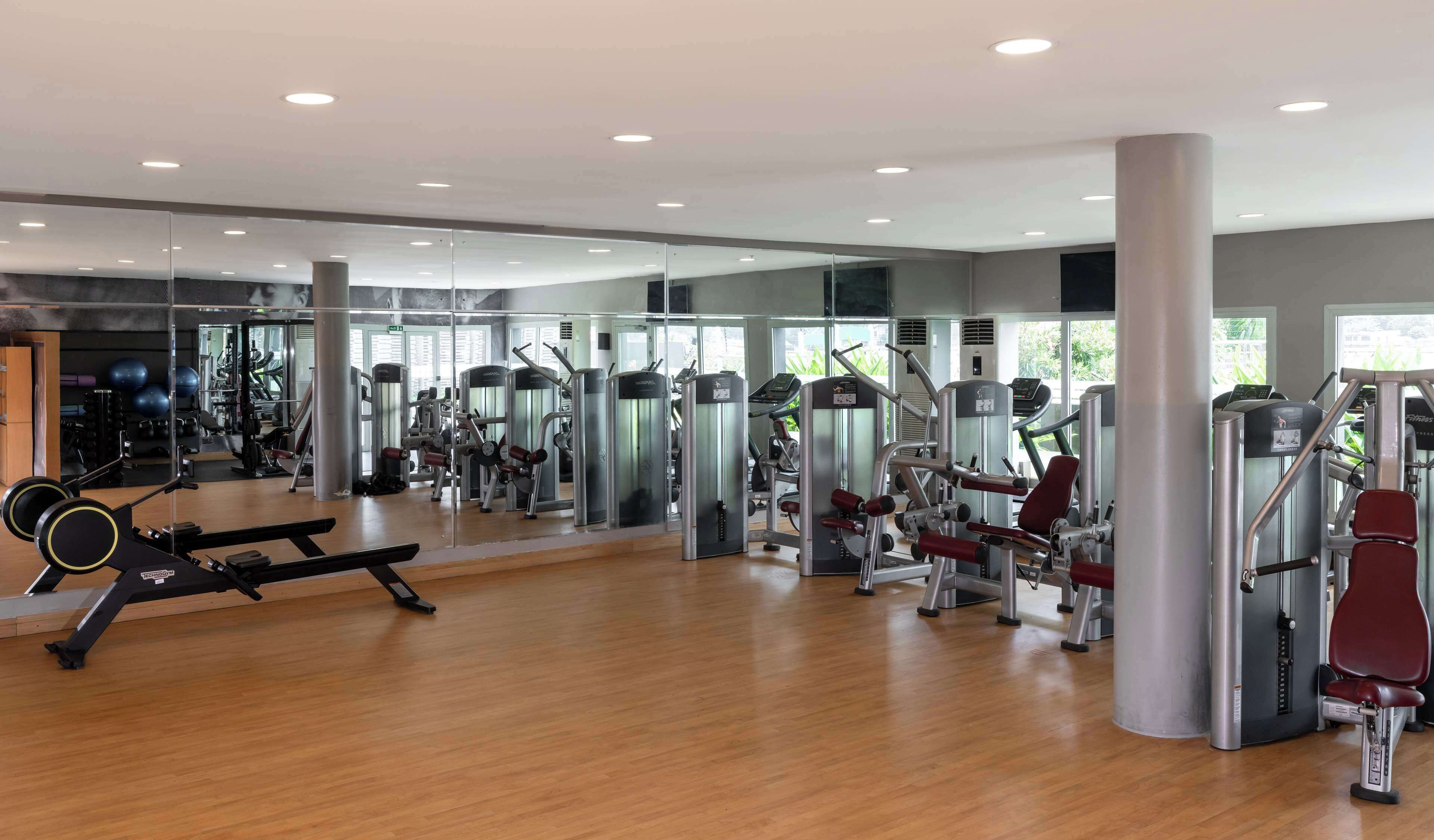 A modern gym with various exercise machines, a mirrored wall, and large windows allowing natural light.