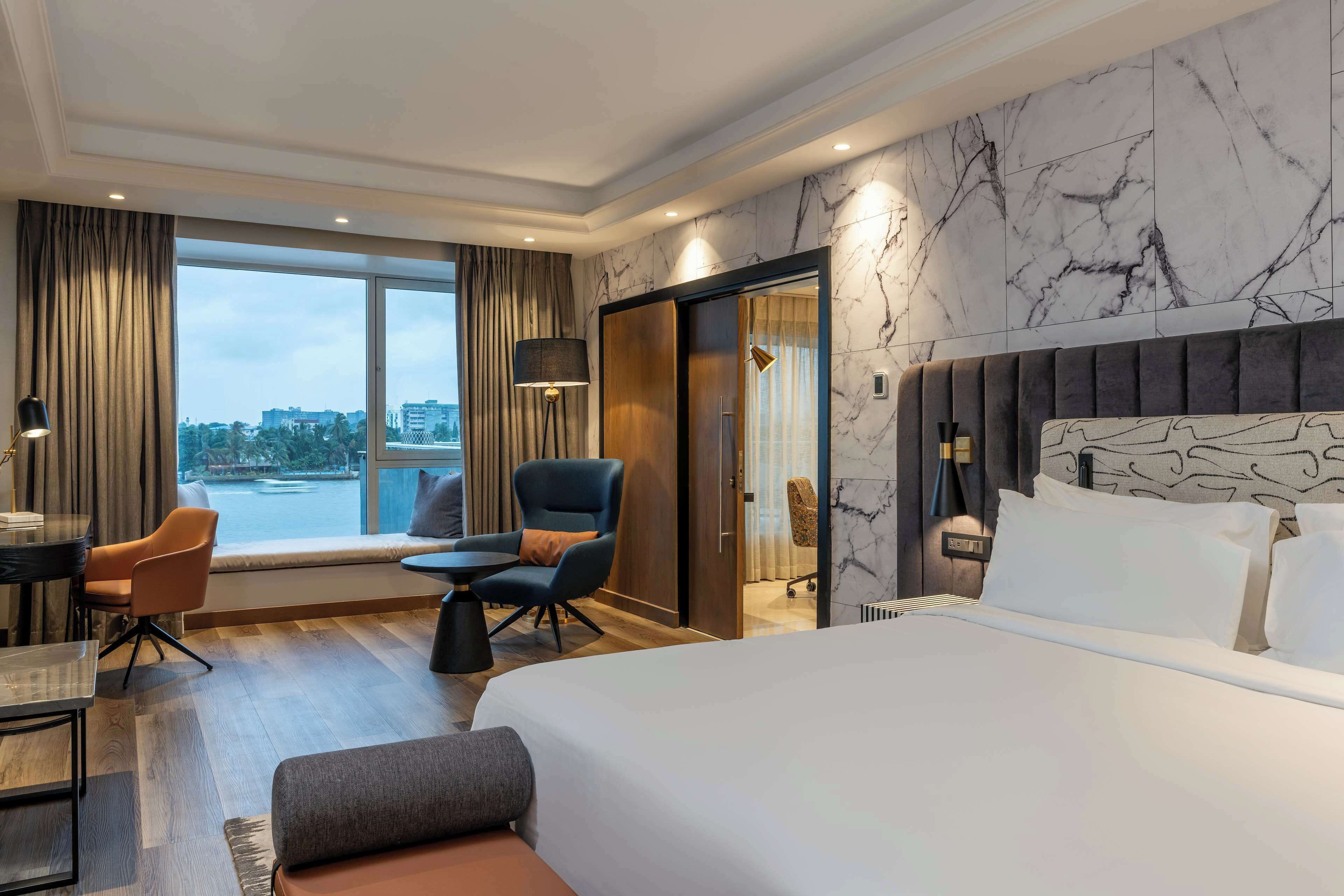 Ein modernes Hotelzimmer mit einem großen Bett, einem Schreibtisch, einem Sessel und Blick auf die Stadt durch große Fenster.