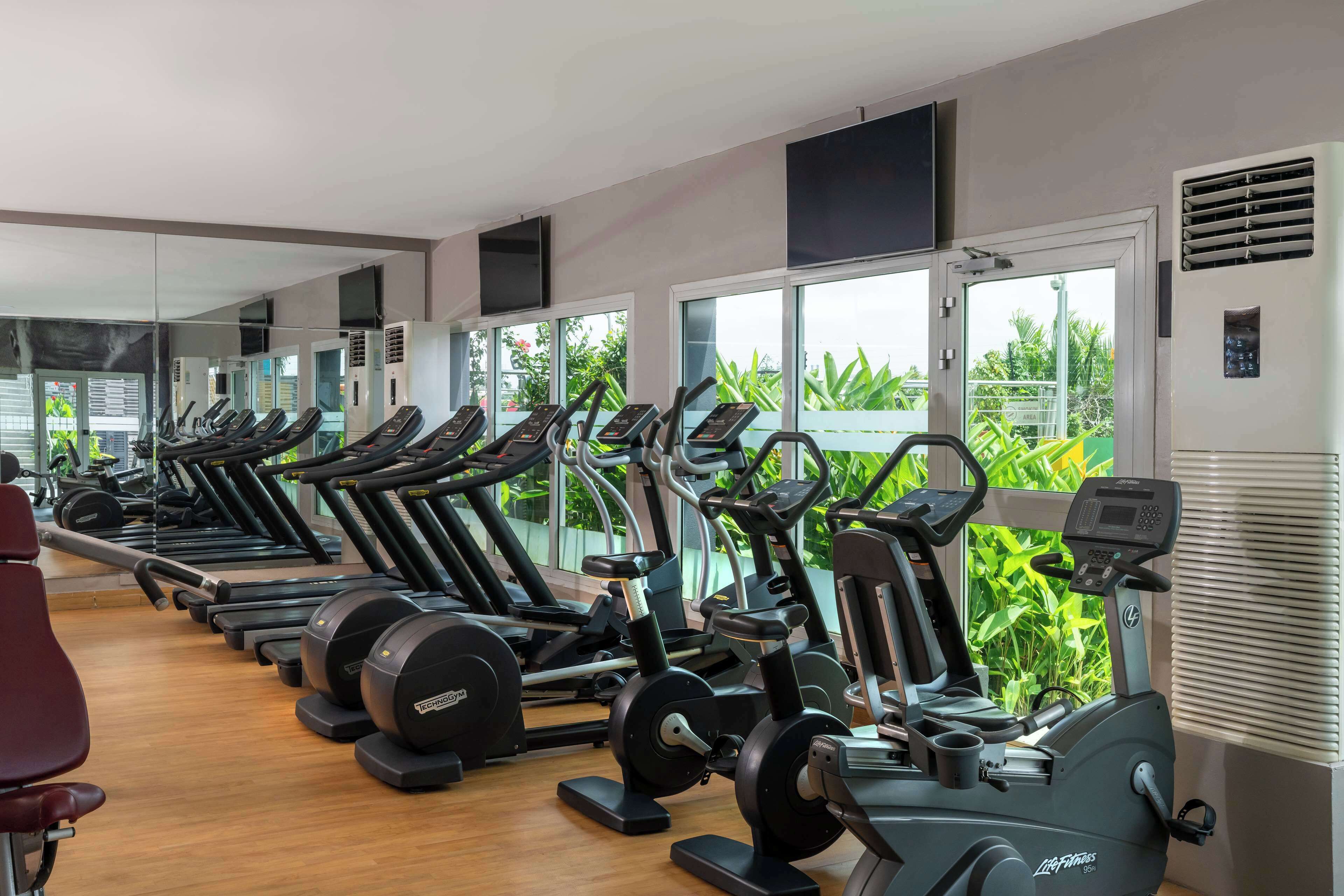 A gym with multiple treadmills and exercise bikes, large windows with a view of greenery, and a wooden floor.
