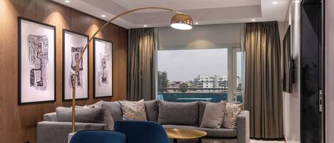 A modern living room with a grey sofa, blue armchair, and a small coffee table. There are framed abstract artworks on the wall and a large window with curtains.