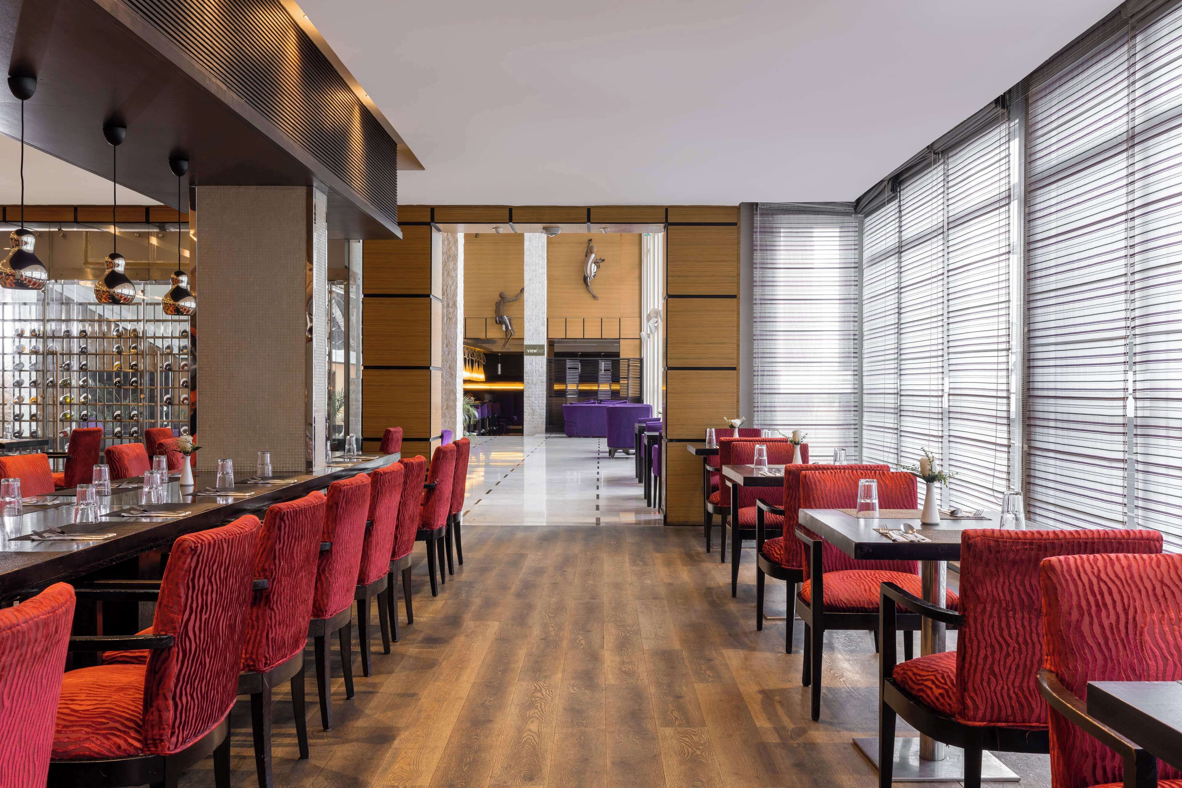 Un ristorante moderno con sedie rosse imbottite, un lungo bancone e vista su un'area bar attraverso una porta in vetro.