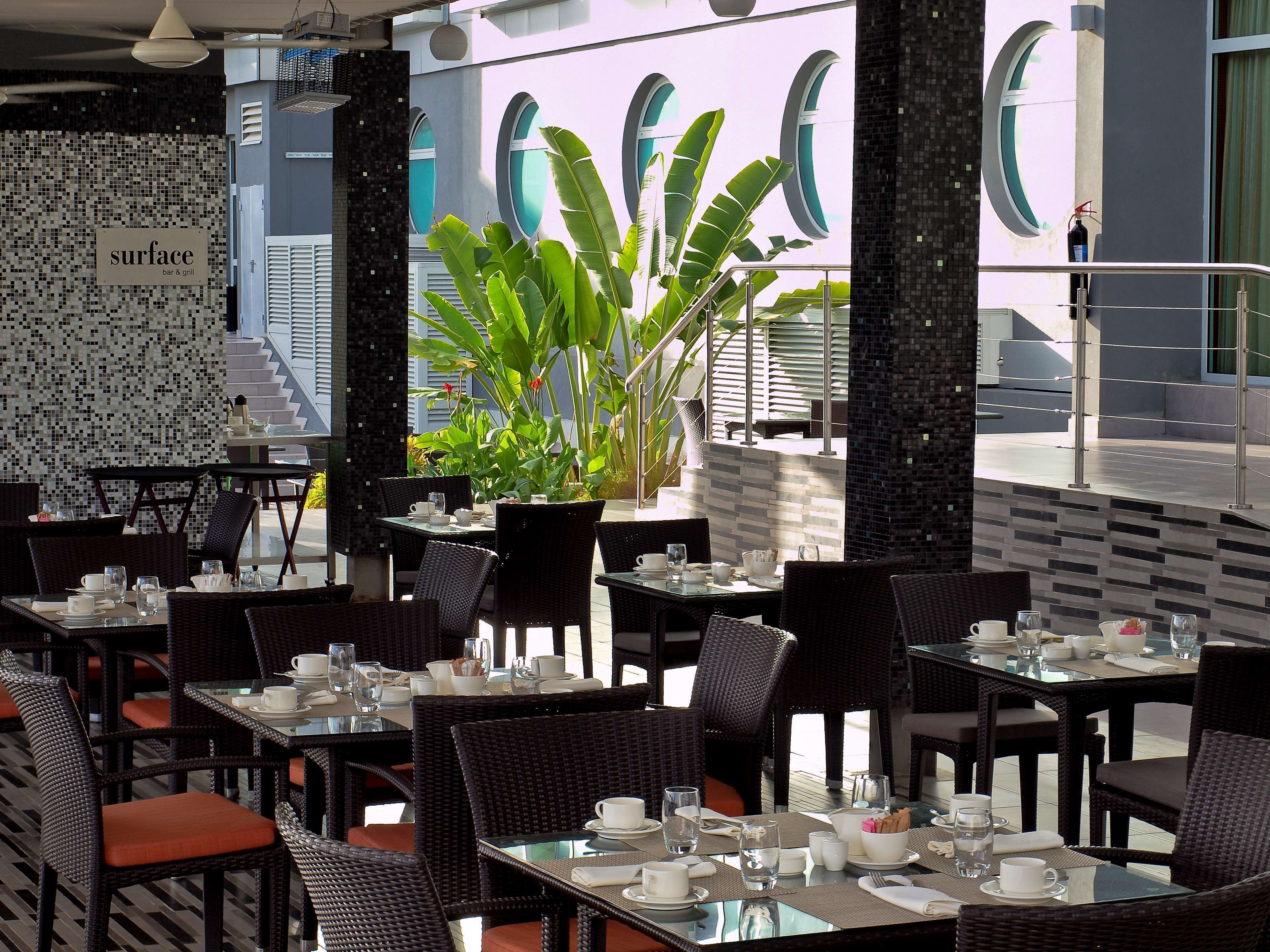A restaurant with glass tables and wicker chairs, a tiled wall with a 'surface bar & grill' sign, and large windows with a view of the street.