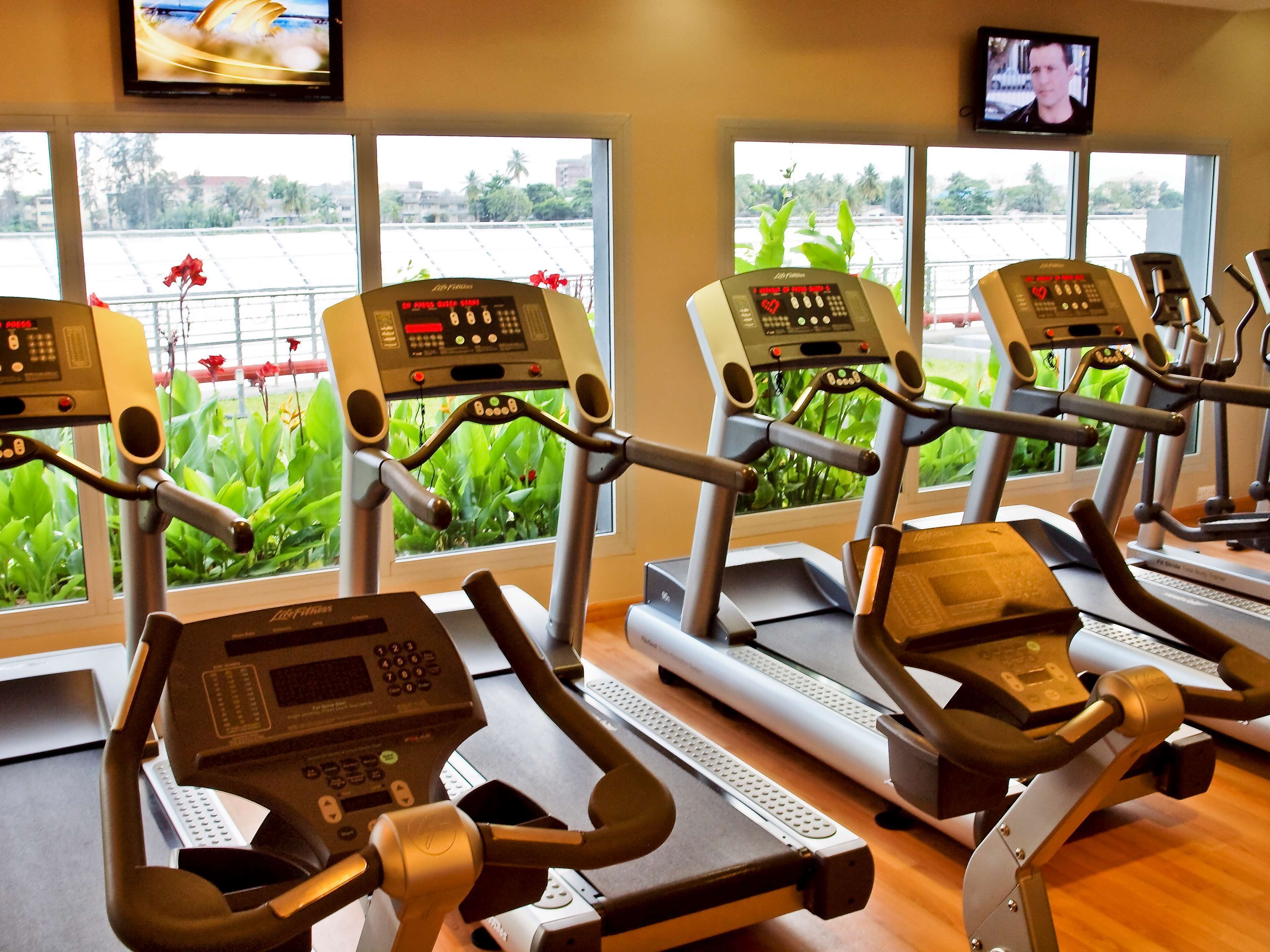 A gym with multiple treadmills, a TV mounted on the wall, and large windows with a view of greenery and a building.