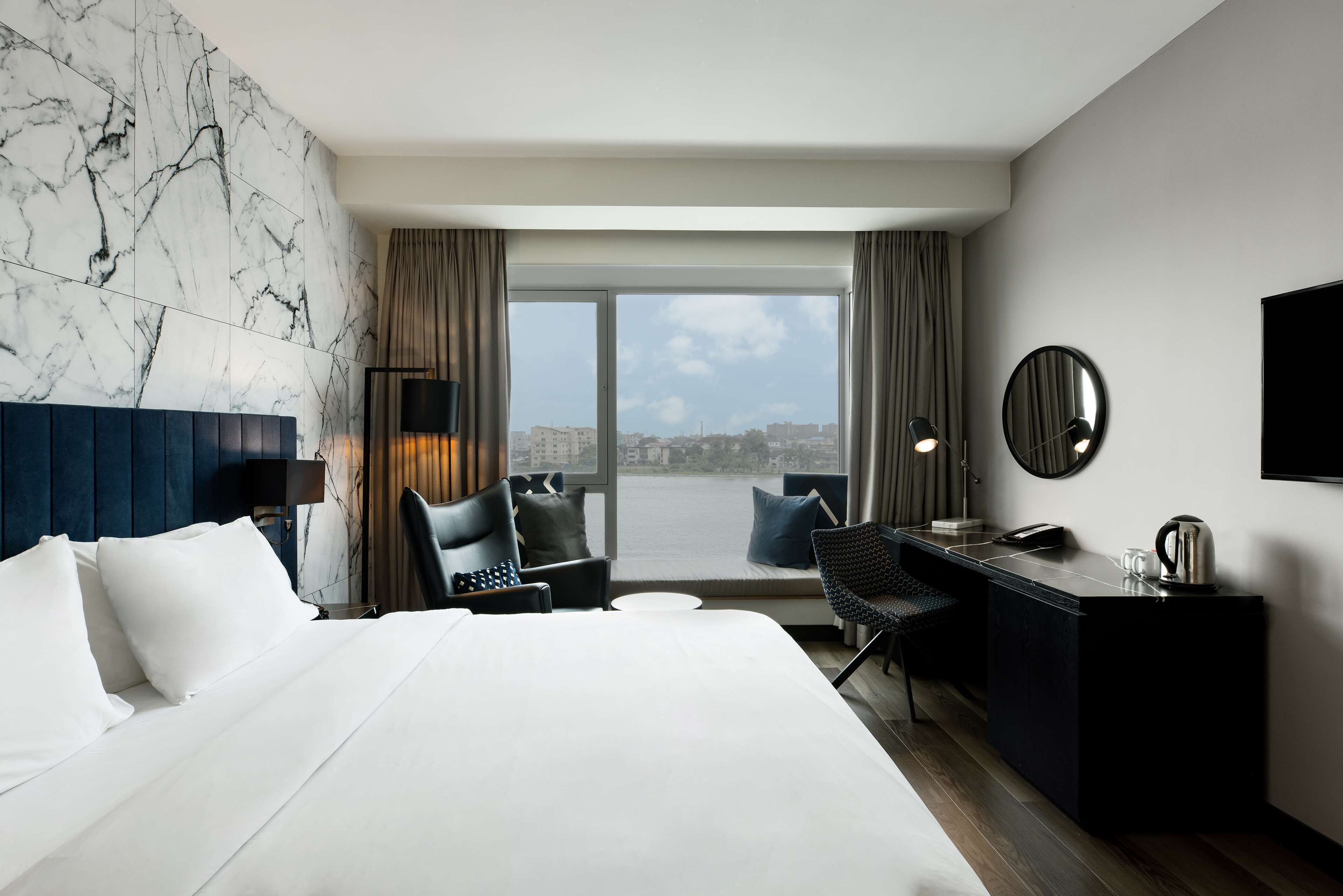 A hotel room with a large bed, a desk, a chair, a small table, a TV, and a window with a city view.