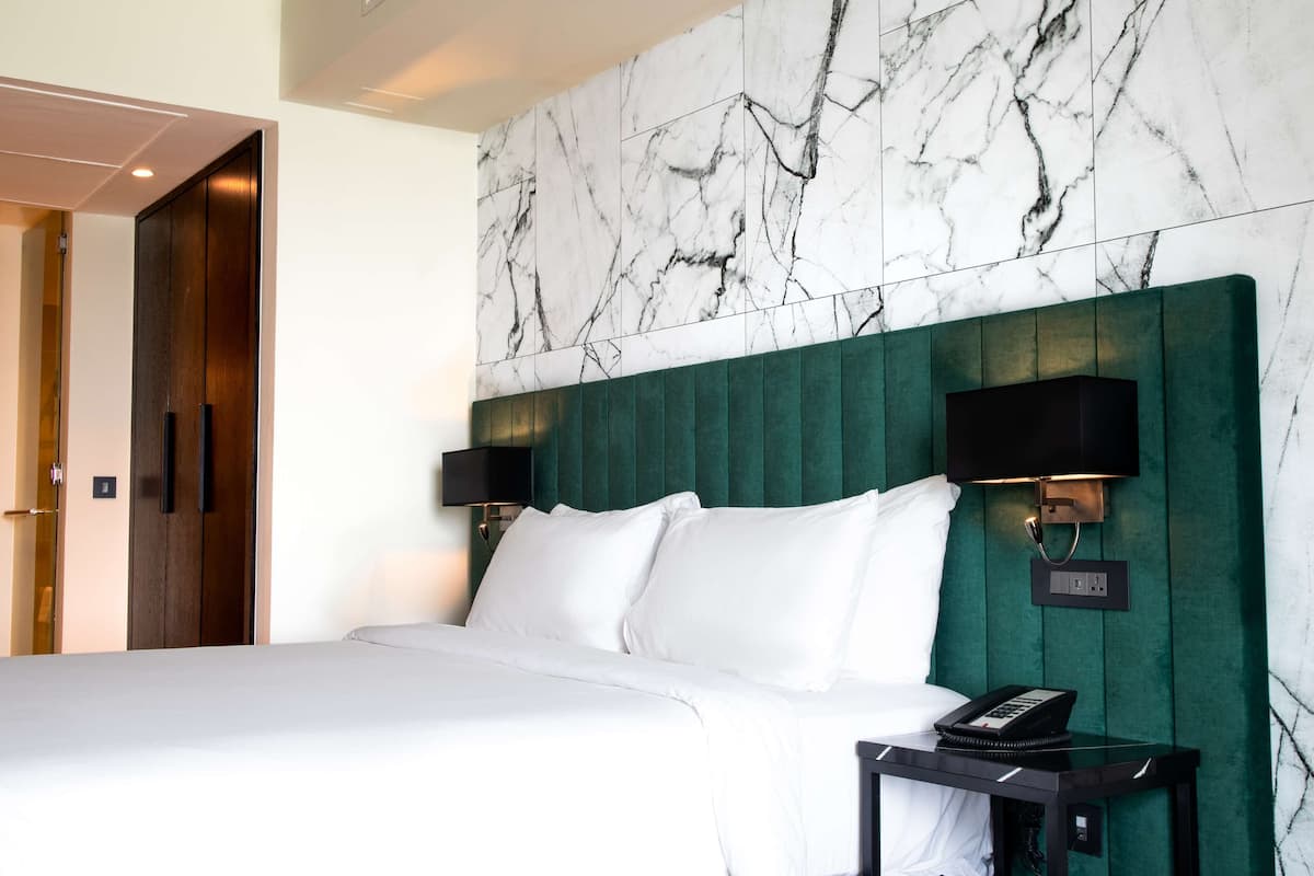A hotel room with a marble-patterned wall, a green upholstered headboard, two bedside lamps, a white bed, and a bedside telephone.