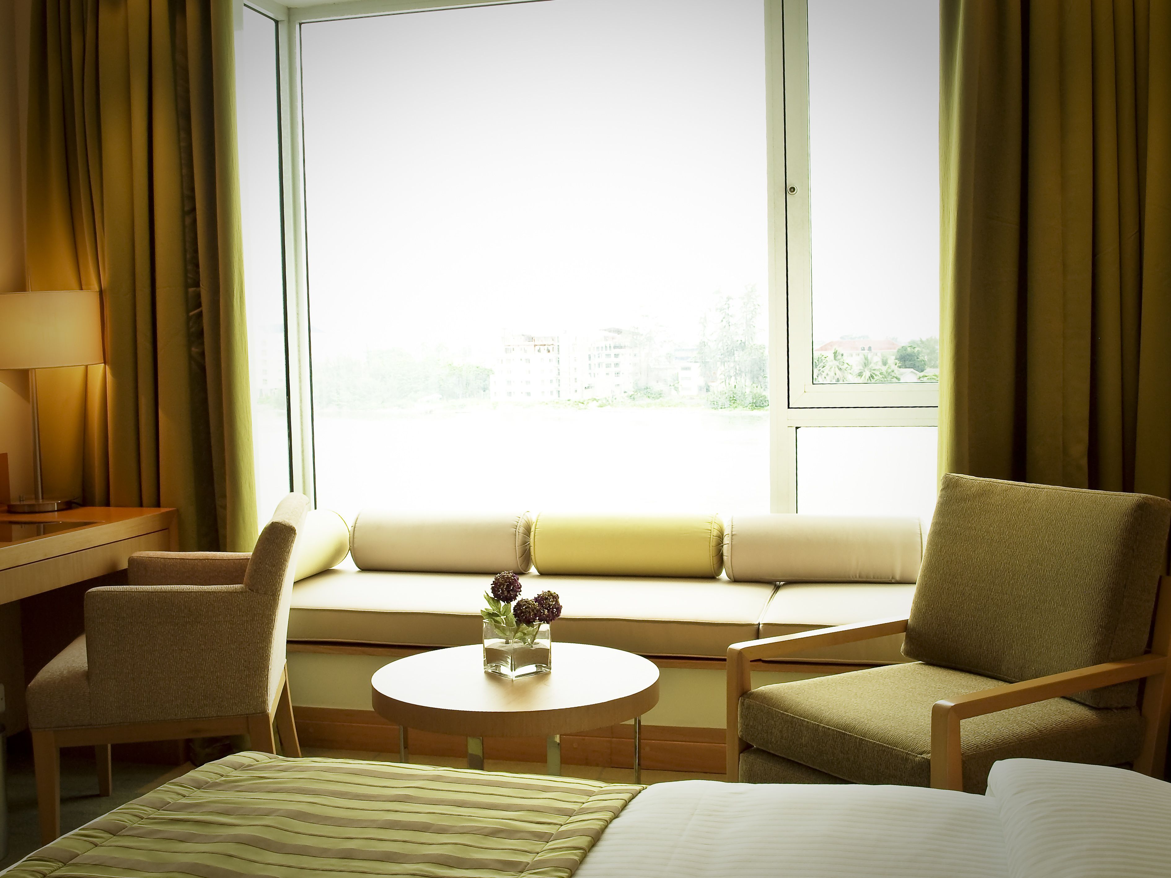 A hotel room with a bed, two armchairs, a small round table with a vase of flowers, and a window with curtains.