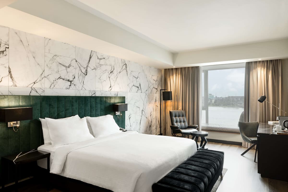 A hotel room with a large bed, a desk, a chair, a view of the water, and marble wall panels.