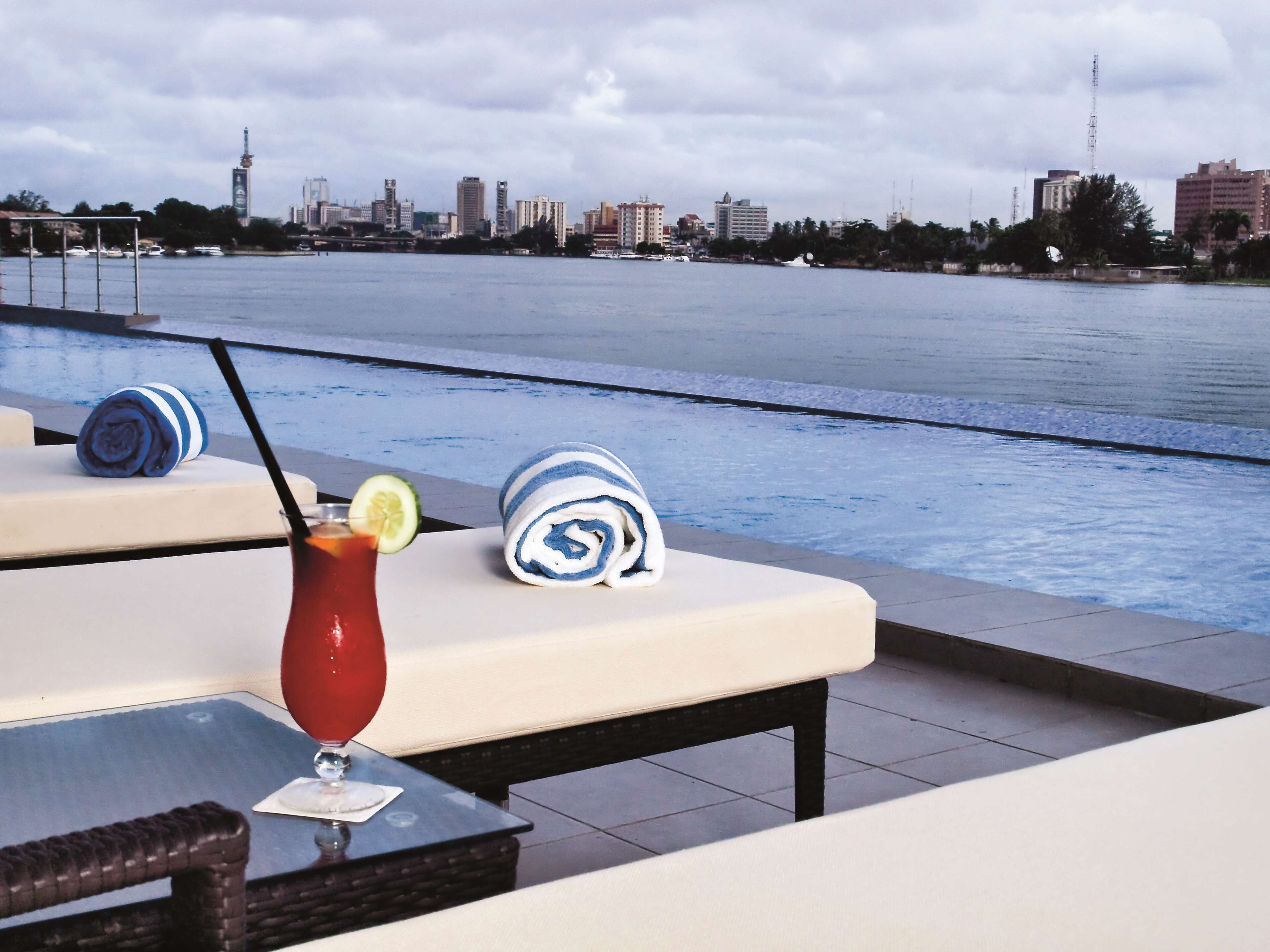 A poolside area with lounge chairs, a table with a drink, and a city skyline in the background.