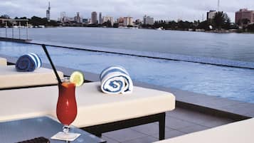 Zona de piscina con sillones reclinables, una mesa con una bebida y un horizonte urbano de fondo.