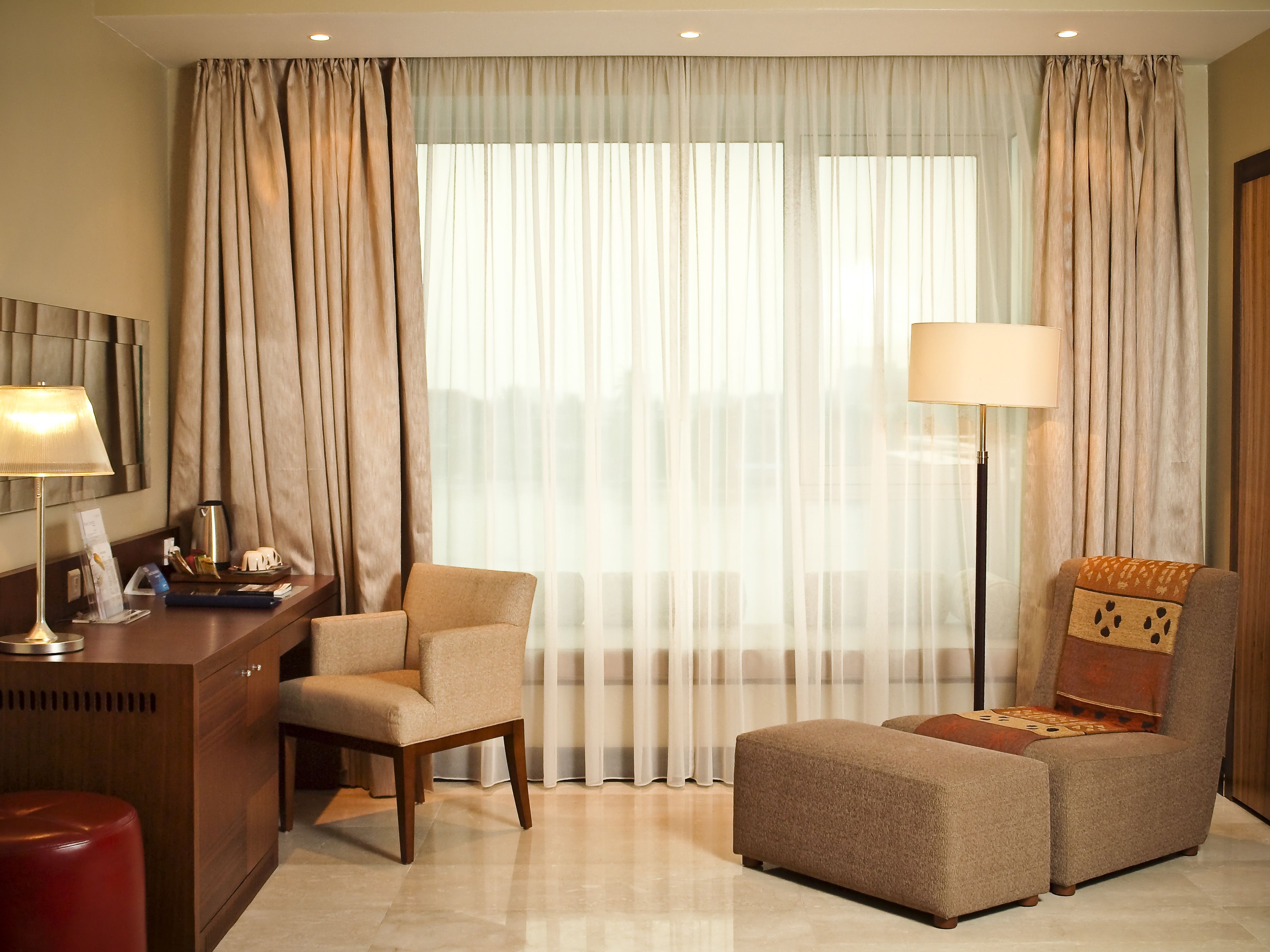 A hotel room with a desk, two armchairs, a red stool, a lamp, and a large window with sheer curtains.