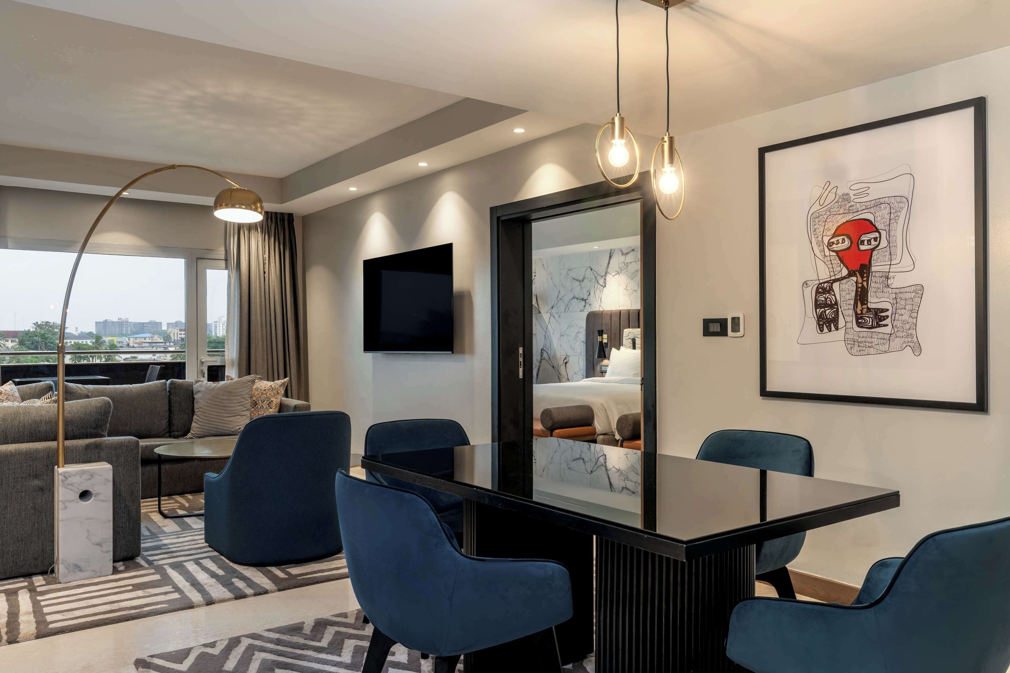 A modern hotel room with a sofa, a dining table, blue chairs, a large mirror, and artwork on the wall.