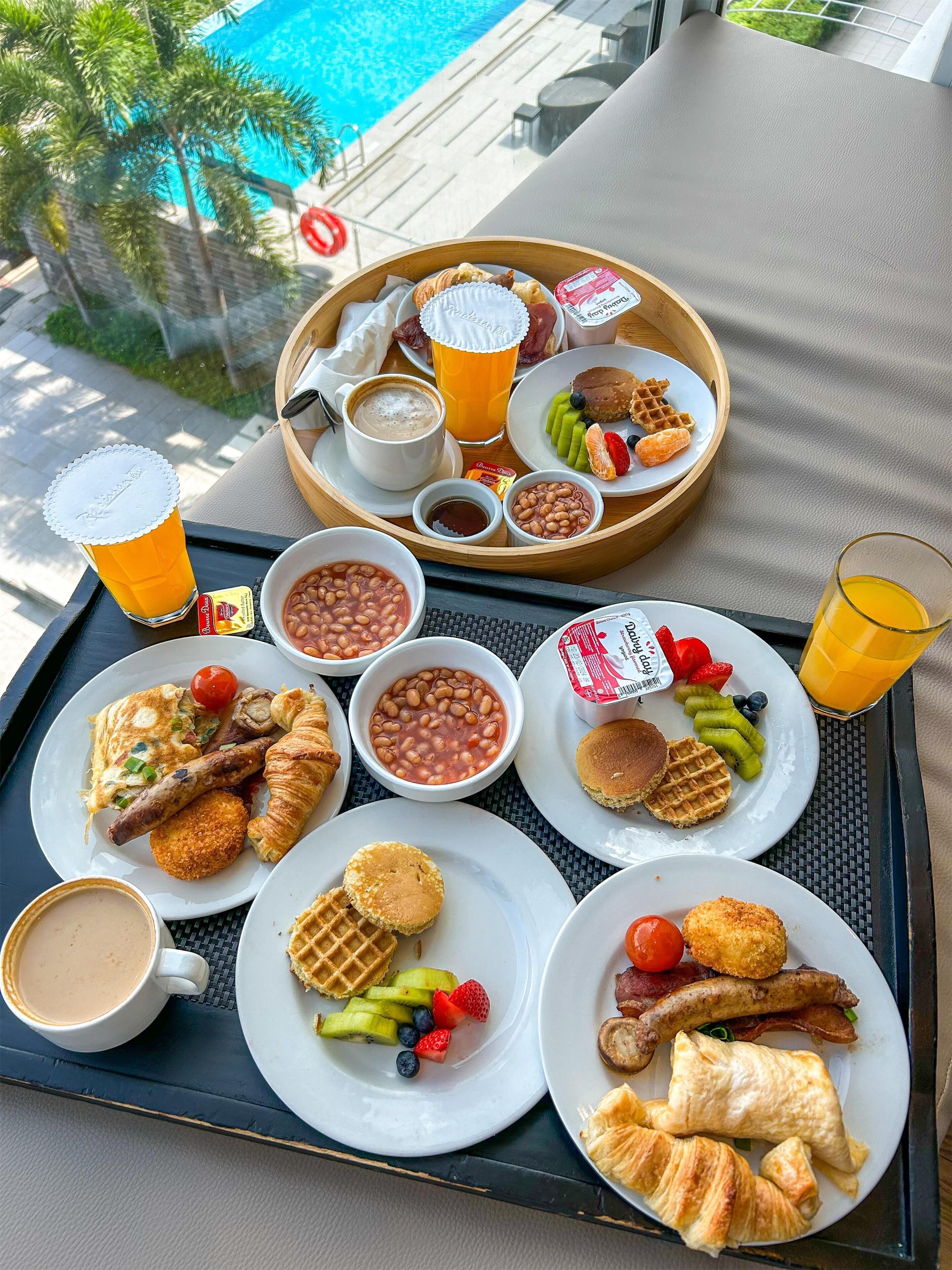 Bandeja de desayuno con diversos alimentos y bebidas, incluyendo gofres, croissants, frijoles y un vaso de jugo de naranja, en un balcón con vista a una piscina.