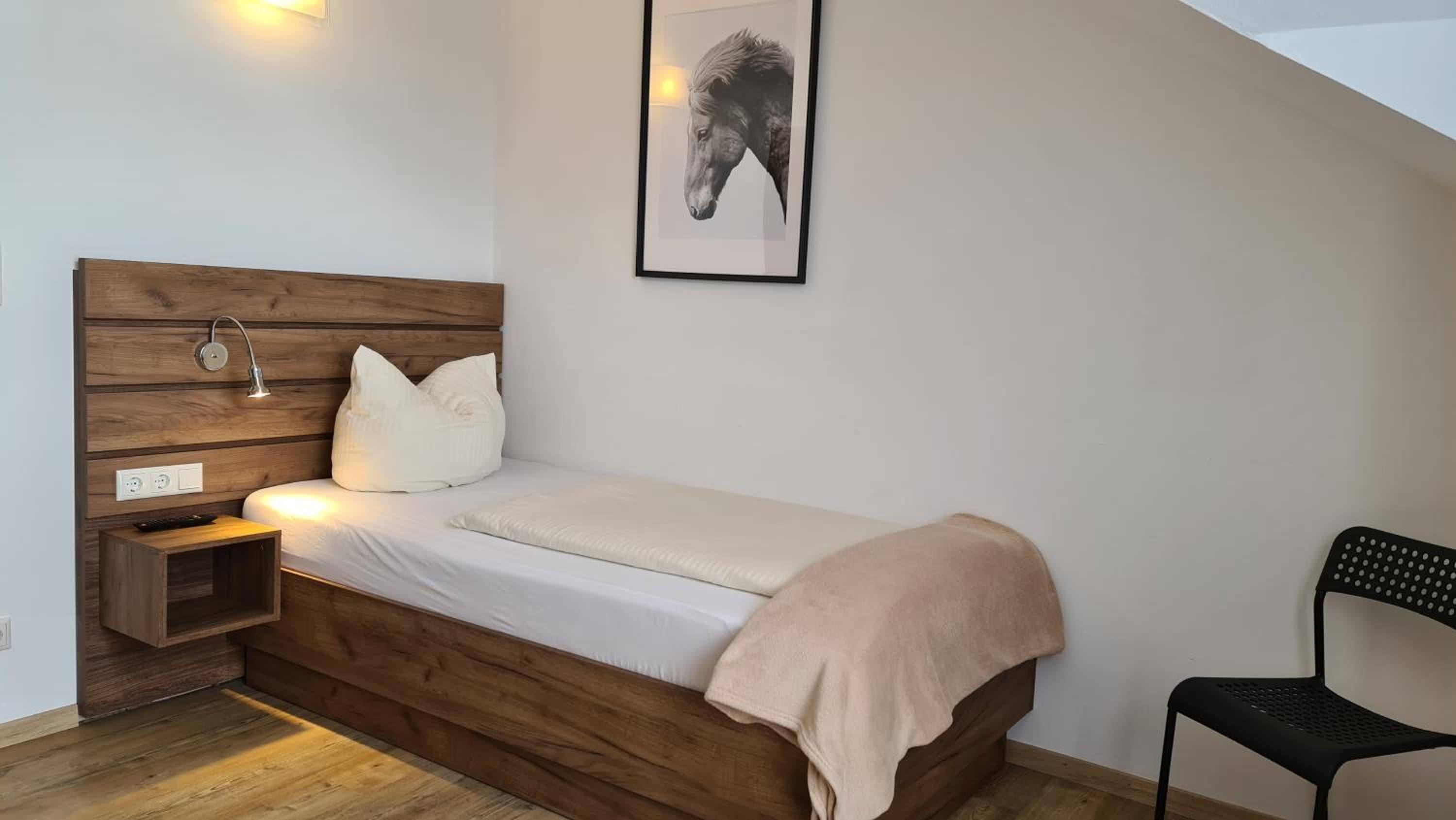 Ein Einzelbett mit Holz Kopfteil, ein Nachttisch mit Lampe, ein Sessel und ein gerahmtes Bild an der Wand.