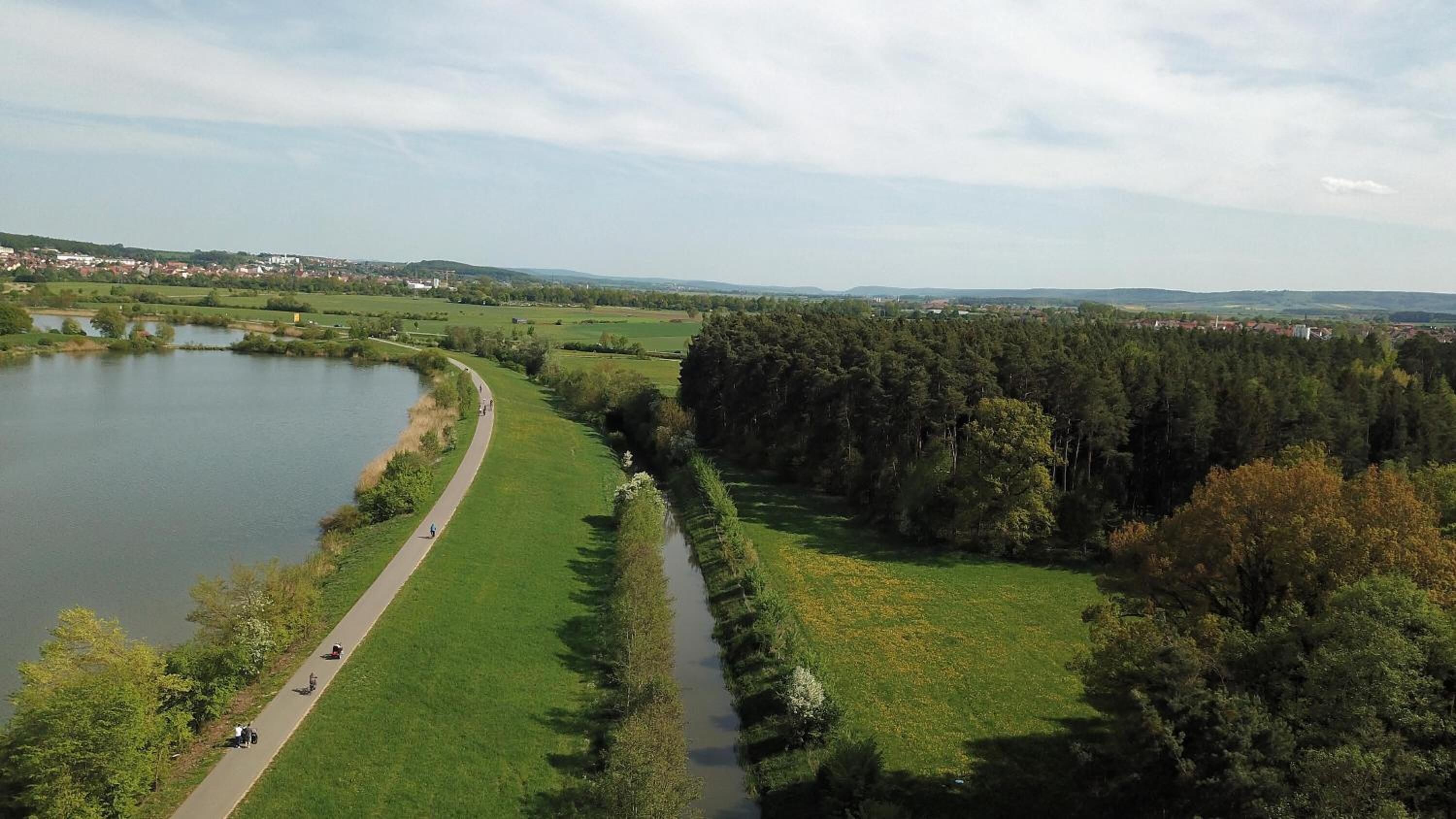 A riverside path with a small bridge, surrounded by greenery and a distant town.