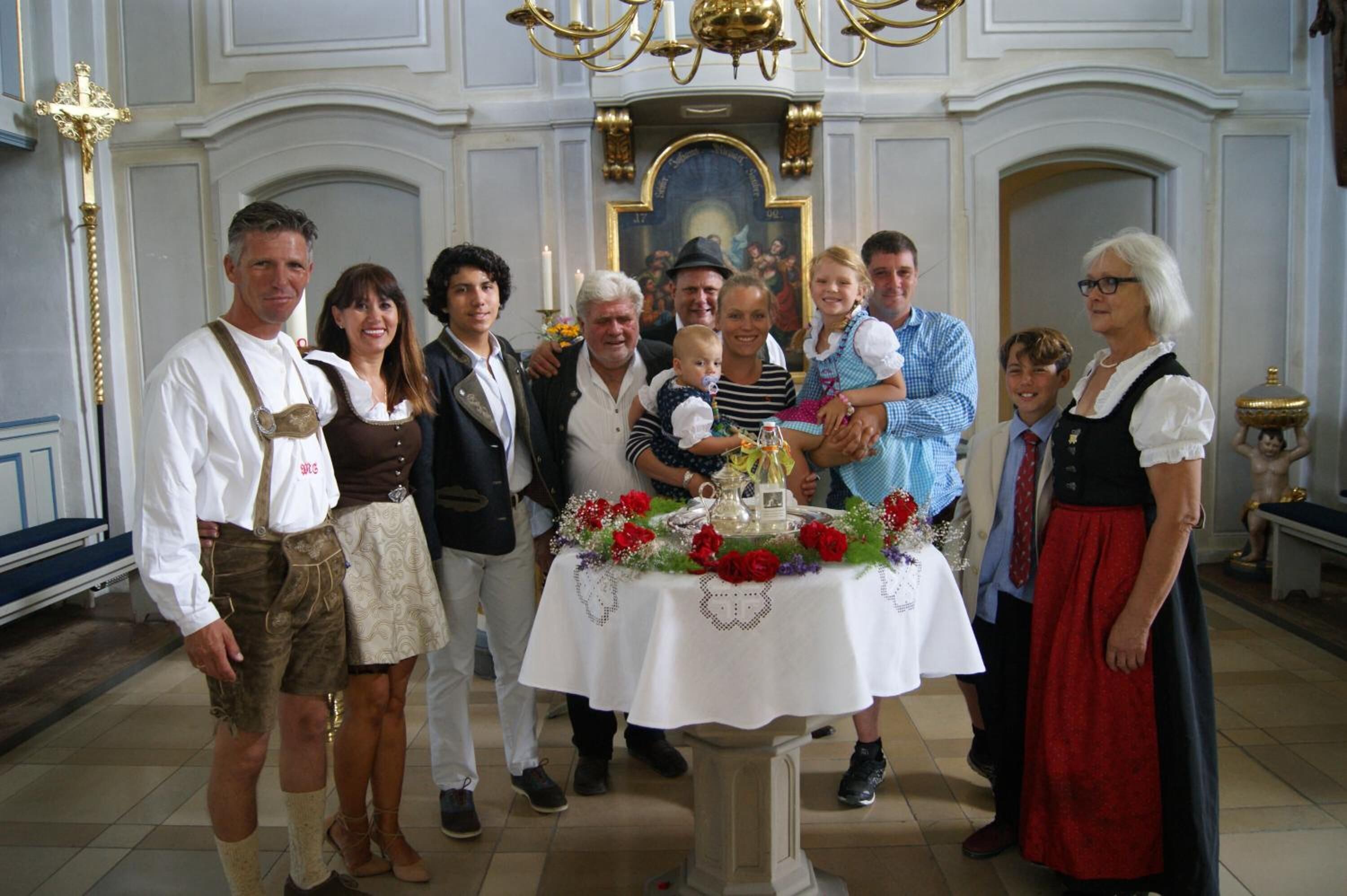 A group of people in traditional attire standing in a room with a table decorated with flowers and a religious painting in the background.