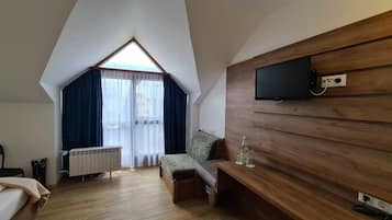 A room with a sloped ceiling, a bed, a bench, a TV mounted on a wooden wall, and a window with curtains.
