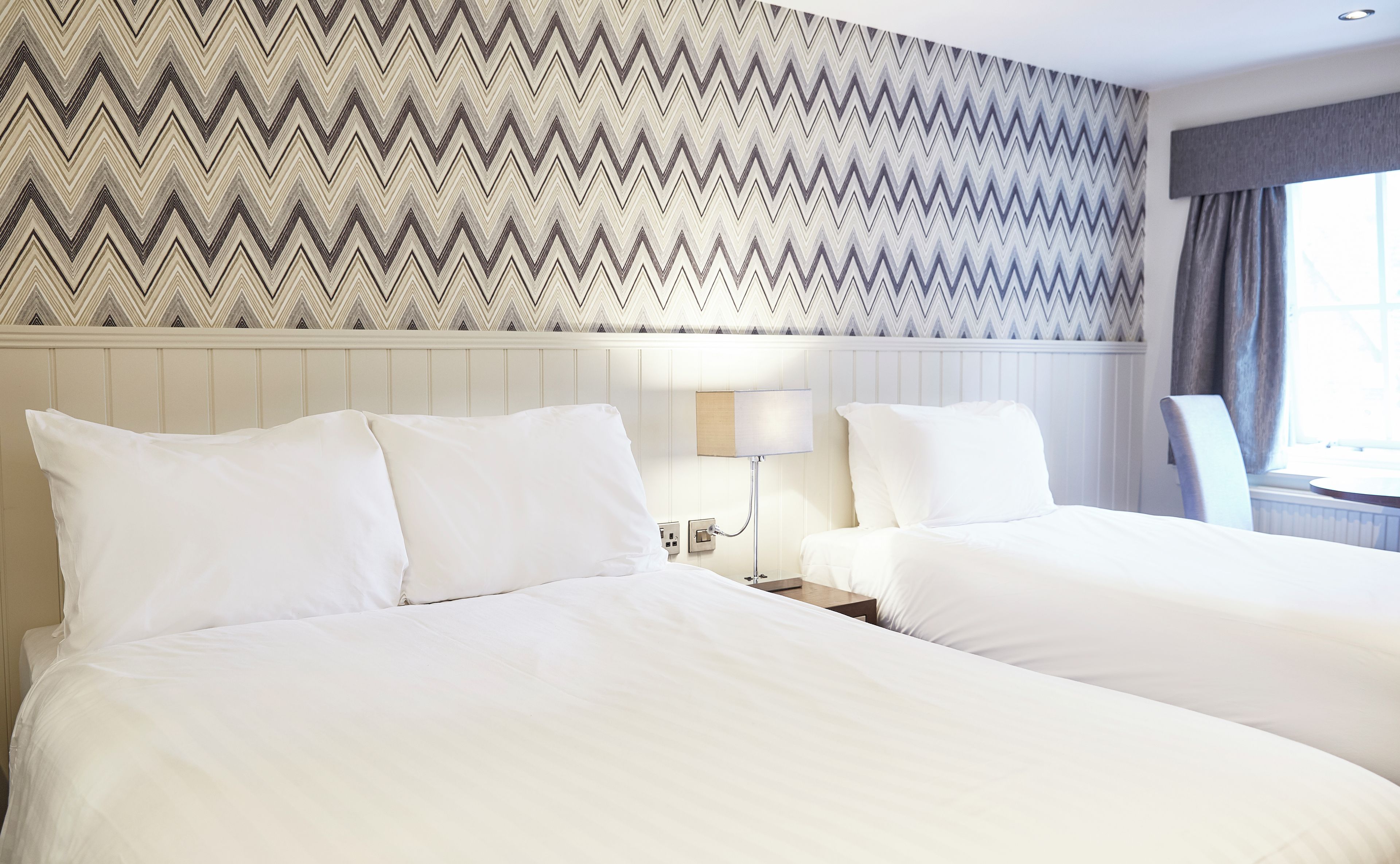 A hotel room with two beds, a nightstand, a lamp, and a window with curtains.