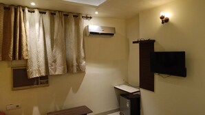 Una habitación con paredes color crema, aire acondicionado instalado, una televisión de pantalla plana, una mesa de madera y una ventana con cortinas.