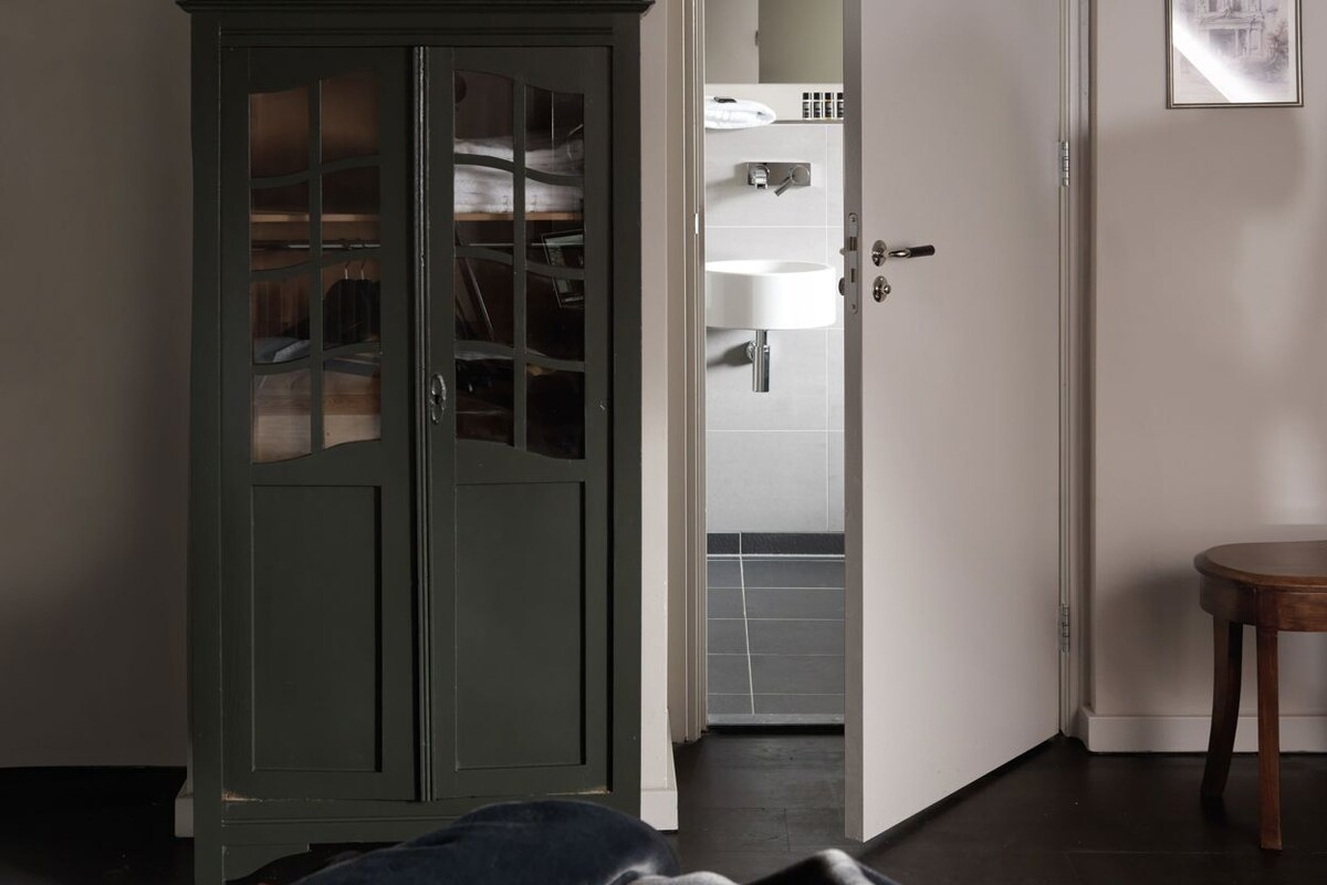 A room with a dark green wardrobe, a bed with a blanket, a small wooden table, and a door leading to a bathroom with a sink and mirror.