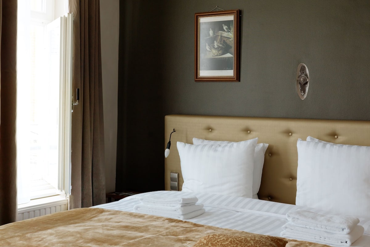 A hotel room with a large bed, a headboard, a framed picture on the wall, and a window with curtains.