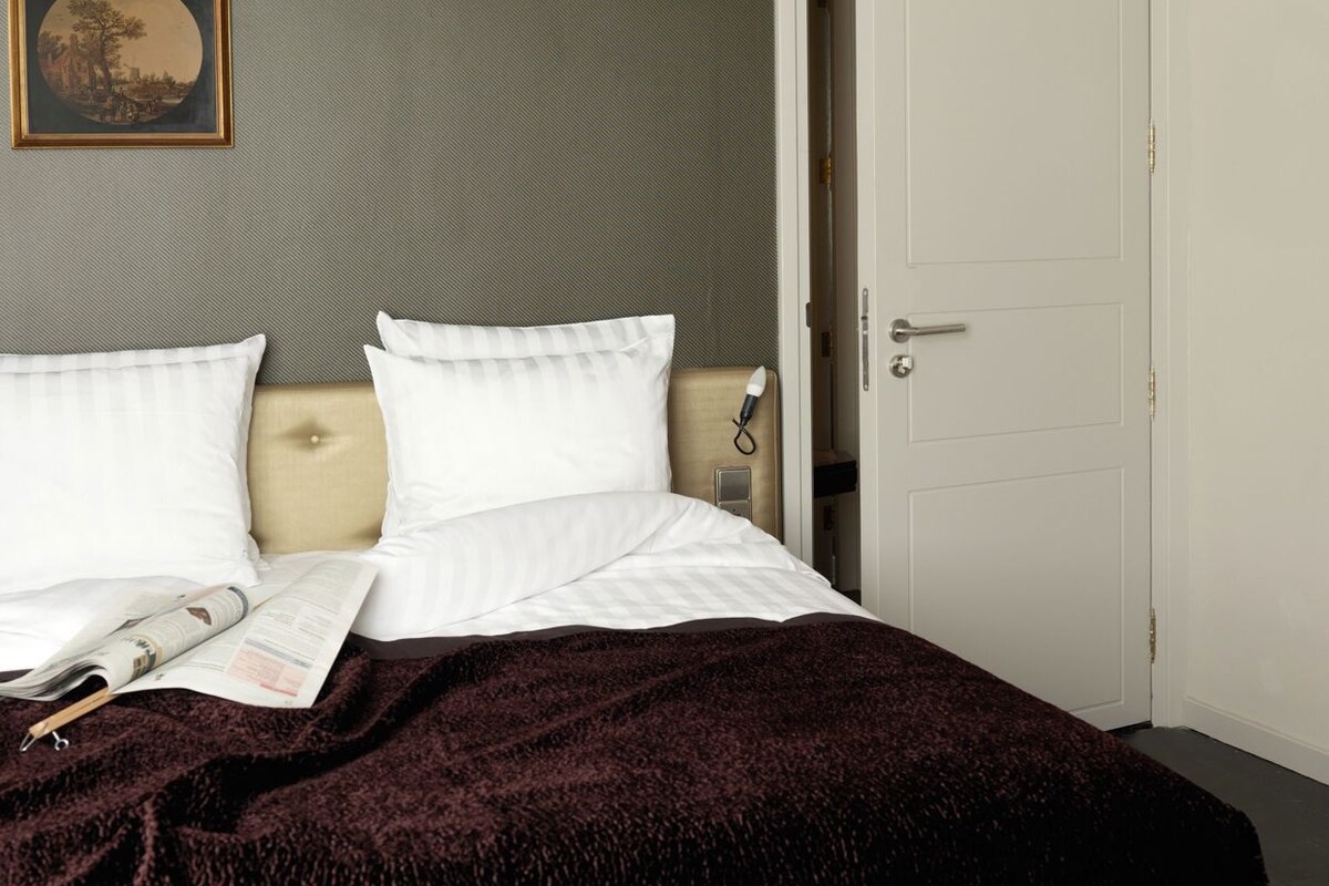 A neatly made bed with a dark blanket, a framed picture on the wall, and a closed door.
