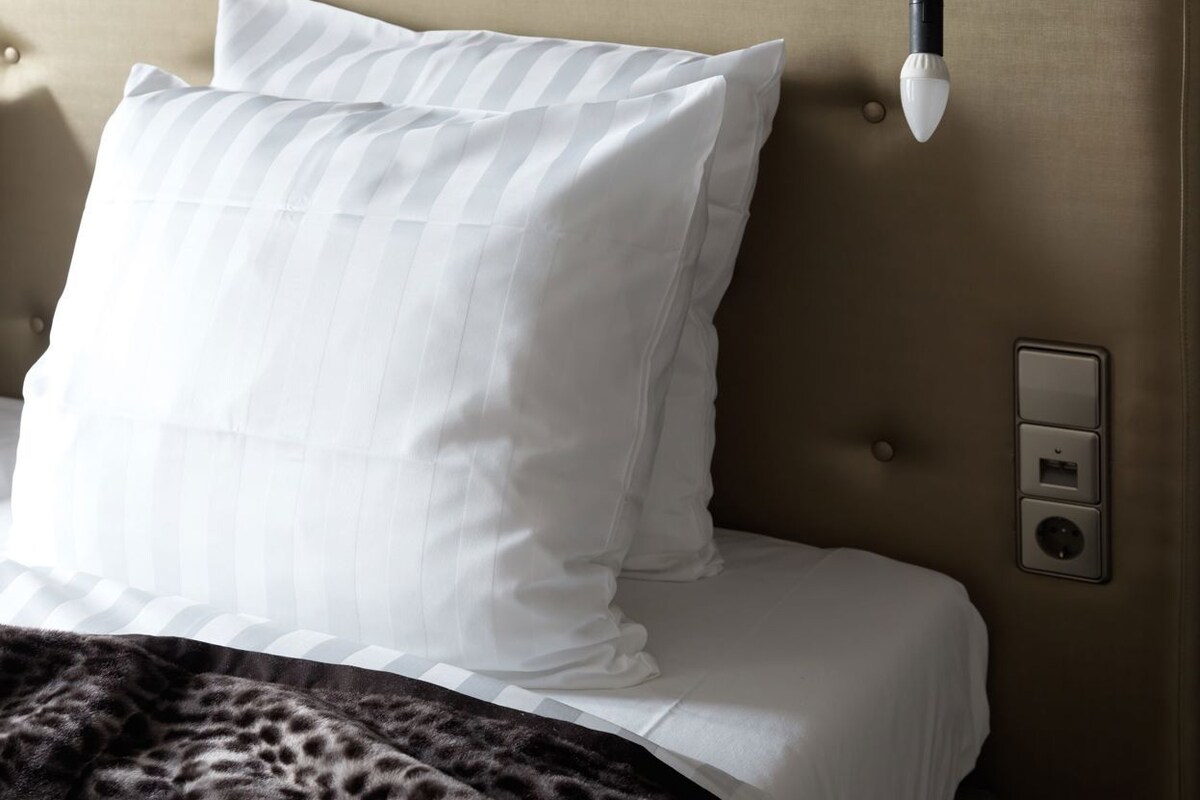 Ein Bett mit einem karriertem Kopfteil, einem weißen Kopfkissen, einer Decke mit Leopardenmuster und einer Wandleuchte.