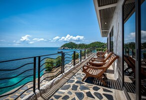 Ein Balkon mit Steinböden, Holzstühlen und einem Geländer mit Blick auf das klare blaue Meer und eine grüne Insel.