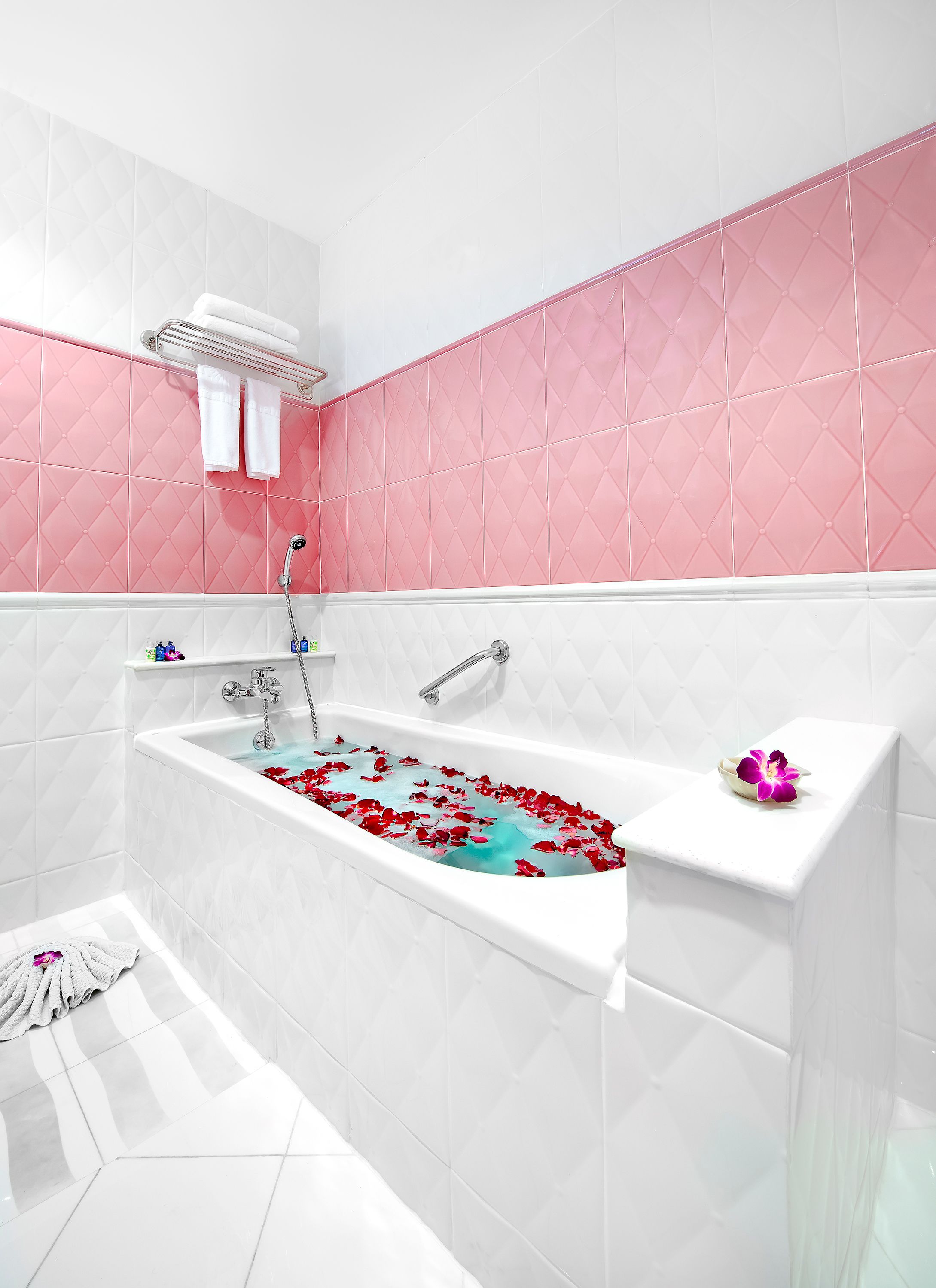 Un baño moderno con una bañera llena de pétalos de rosa, paredes cubiertas de azulejos blancos y rosados, y un lavamanos blanco con una orquídea decorativa.