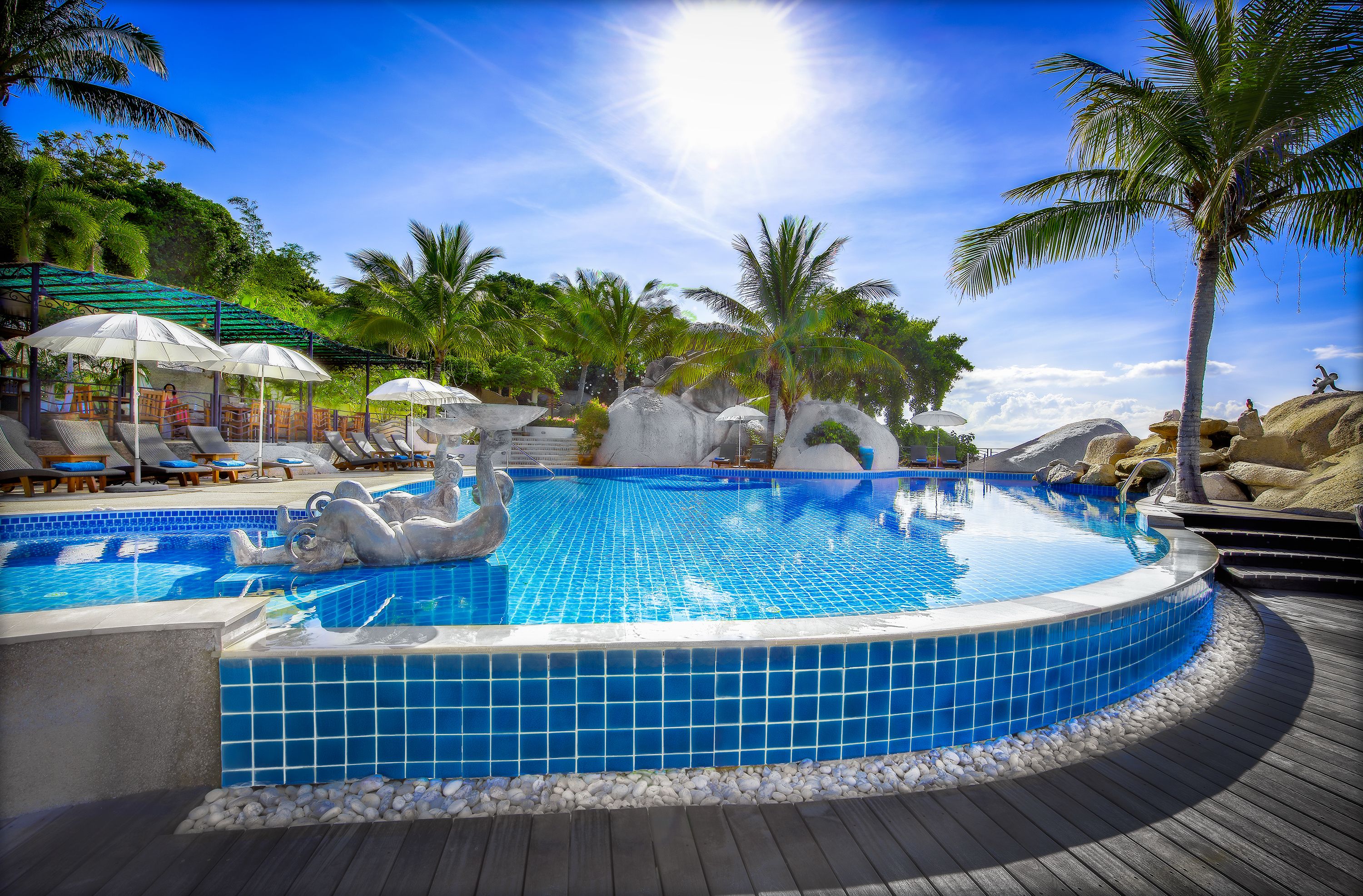 A swimming pool with a decorative swan statue, surrounded by lounge chairs and umbrellas, with palm trees and rocks in the background.