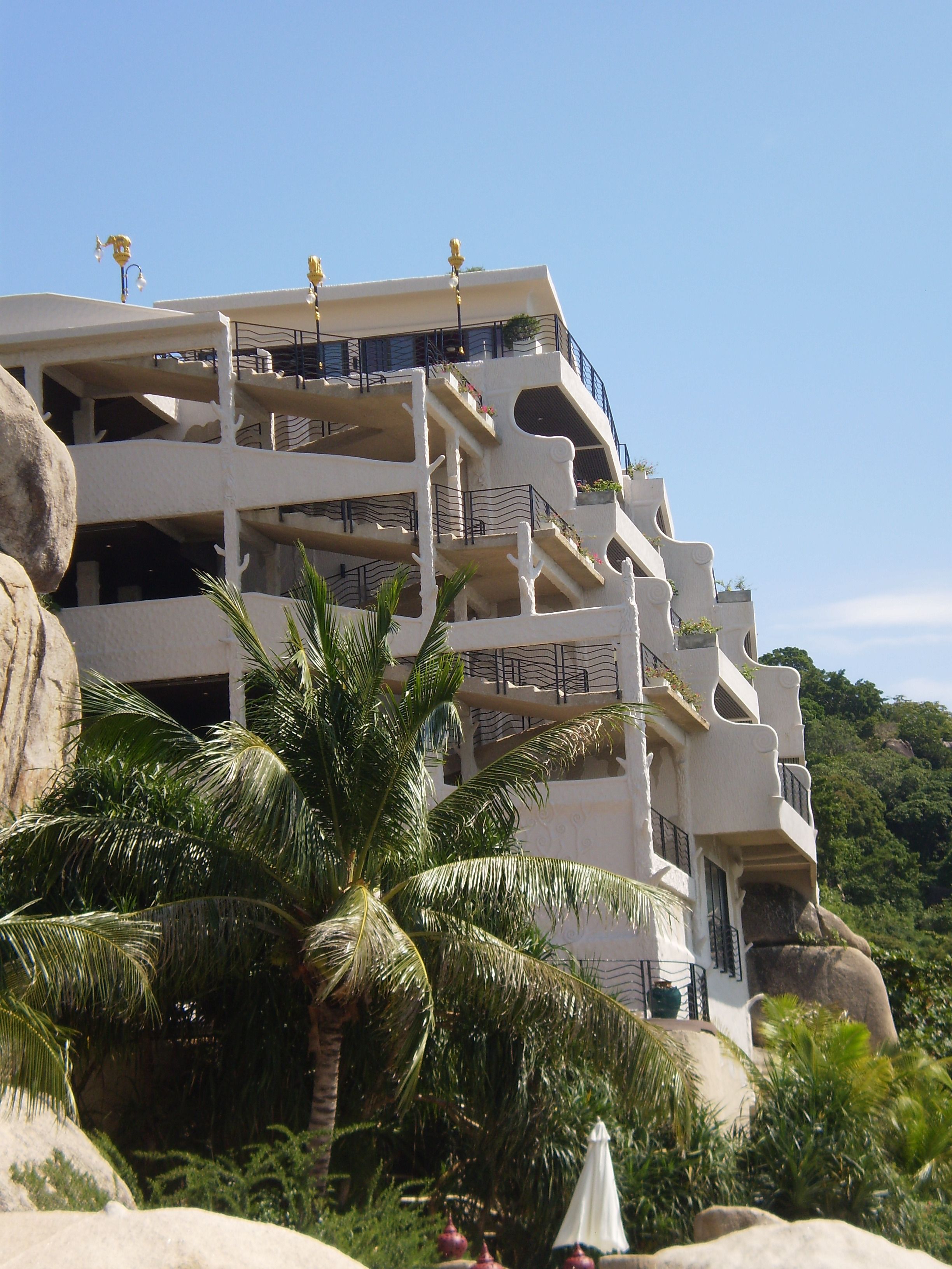 A multi-story building with balconies, surrounded by palm trees and rocks.