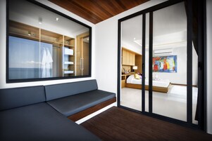 Una habitación moderna con un ventanal grande, un banco integrado y una puerta corredera que da a un dormitorio con una cama y un cuadro colorido.