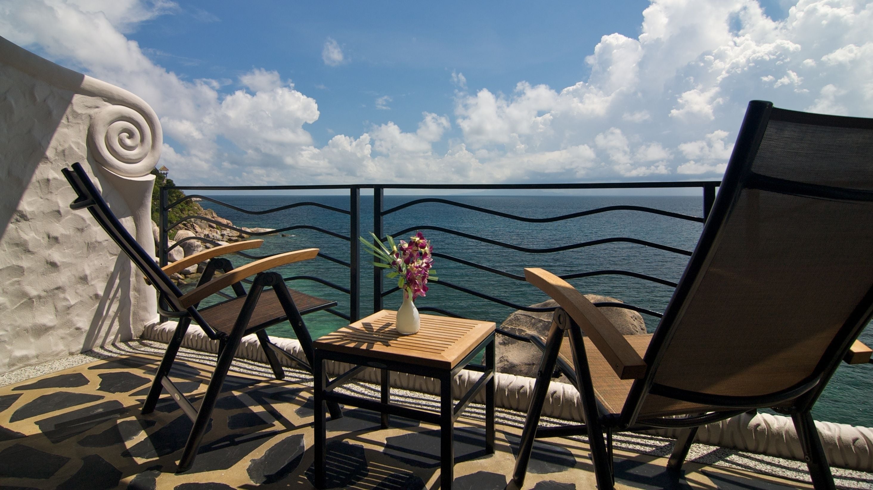 Un balcon donnant sur la mer, aménagé avec deux chaises et une petite table sur laquelle est posé un vase de fleurs.