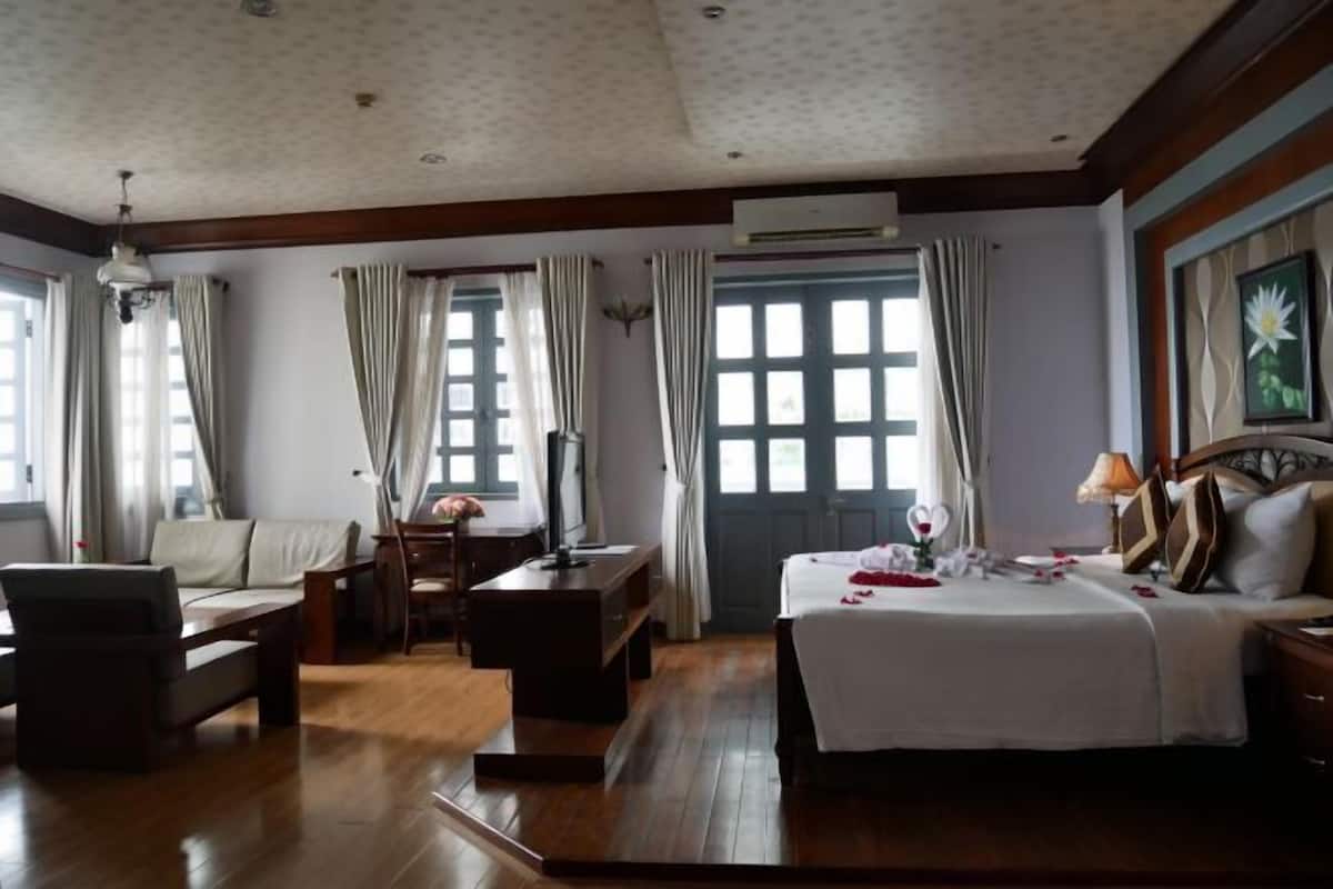 Une chambre d’hôtel avec un lit, un canapé, un bureau et une chaise. Il y a une fenêtre avec des rideaux, un appareil de climatisation et un tableau encadré au mur.