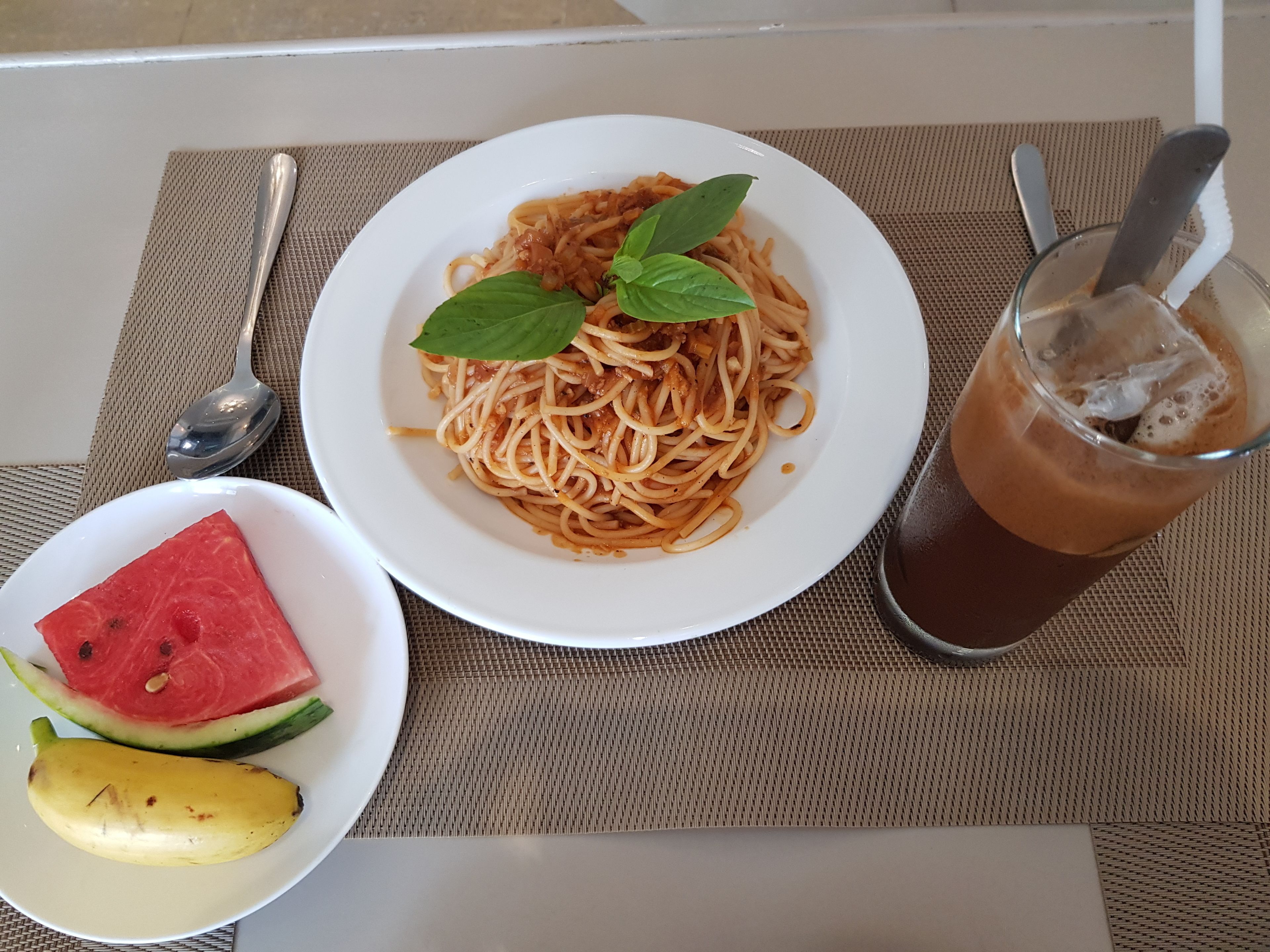 Um prato de espaguete com manjericão, um copo de café gelado, uma fatia de melancia e uma banana sobre uma mesa.