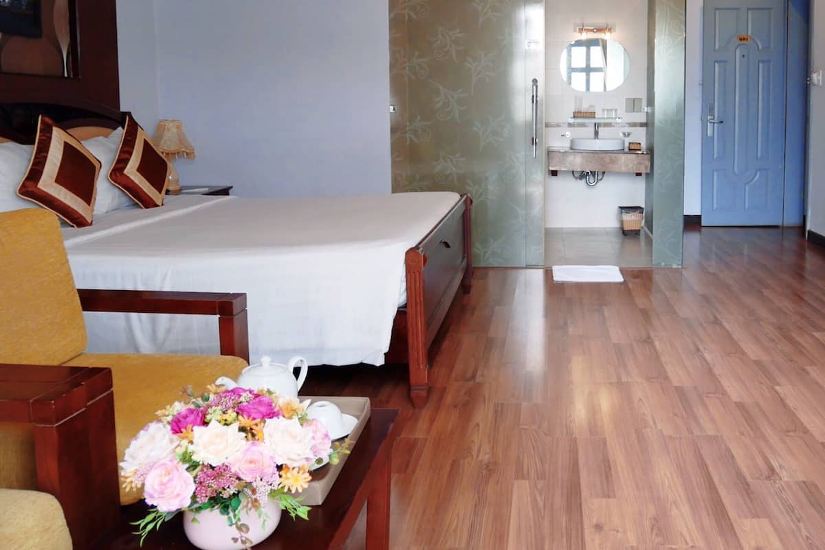 Une chambre d’hôtel avec un lit, un bureau, une chaise, un vase de fleurs et une salle de bain visible à travers une porte en verre.