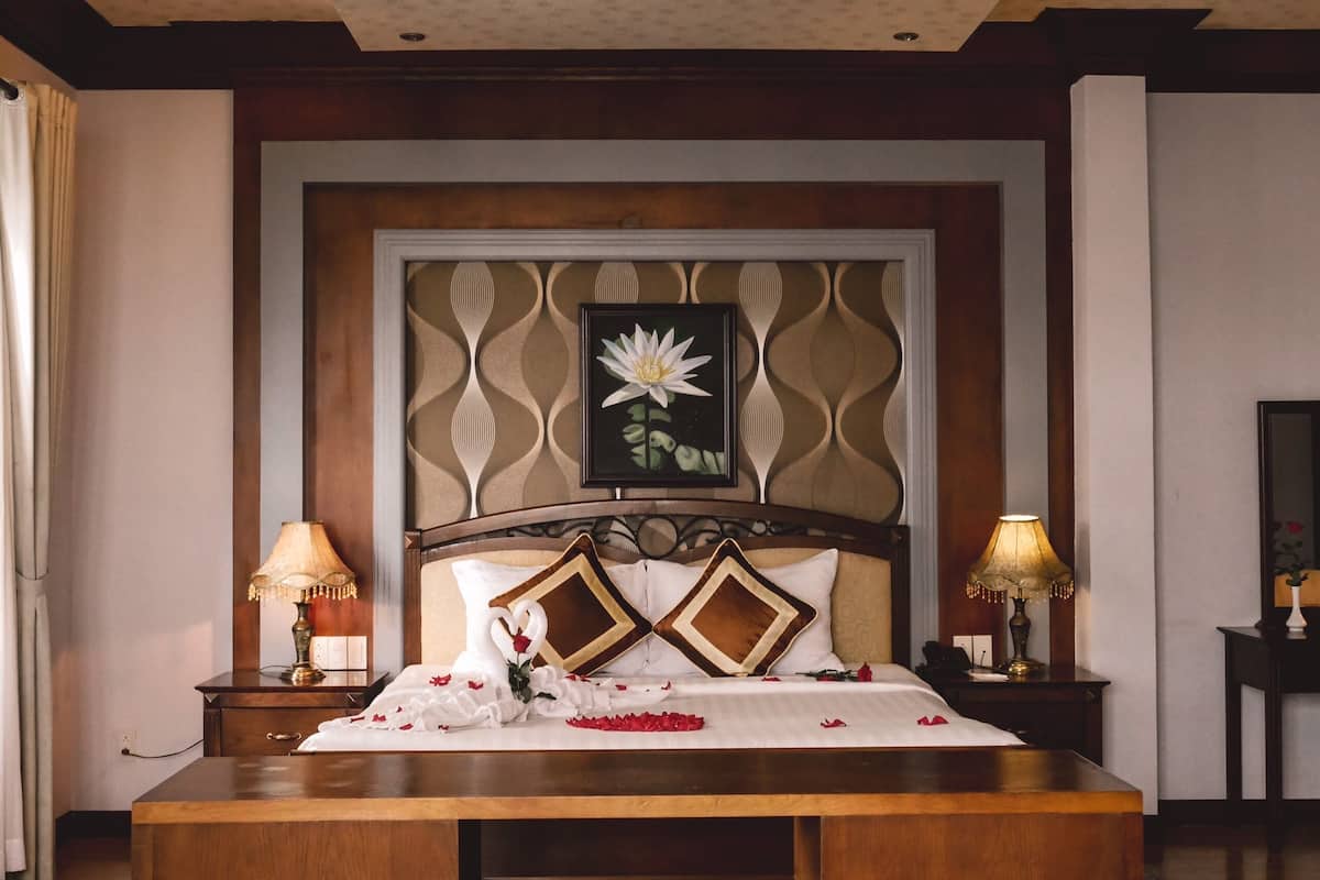Une chambre d’hôtel avec un grand lit, deux tables de chevet avec des lampes, une tête de lit décorative et une œuvre d’art florale encadrée au-dessus du lit.