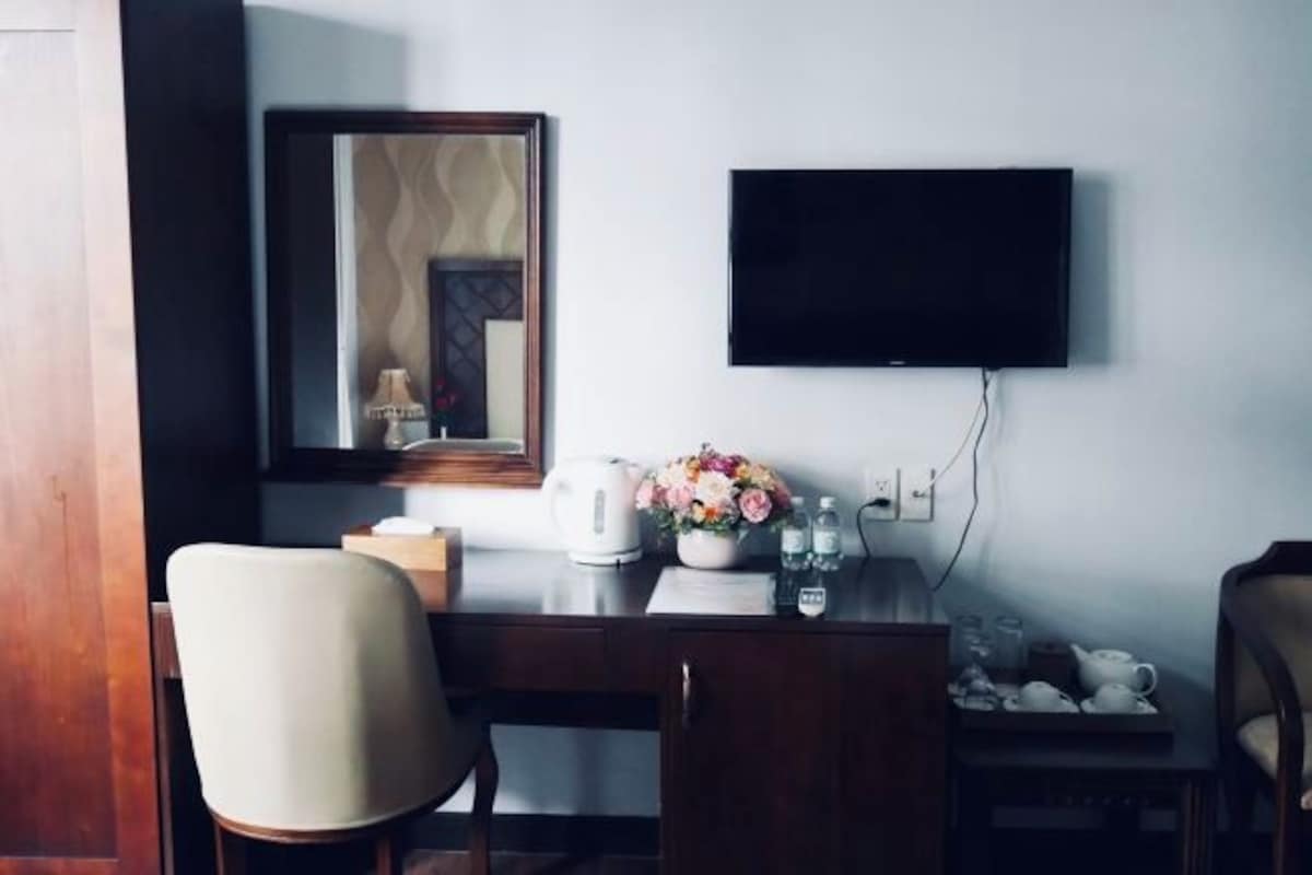 Une chambre d’hôtel équipée d’un bureau, d’une chaise, d’un téléviseur à écran plat, d’un miroir, ainsi que d’une petite table agrémentée d’un vase de fleurs et d’une théière.