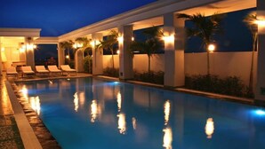 Una piscina amb gandules i palmeres, envoltada per un edifici amb columnes i il·luminació càlida.