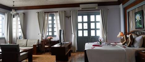 A hotel room with a bed, a sofa, a desk, and a chair. There is a window with curtains, an air conditioning unit, and a painting on the wall.