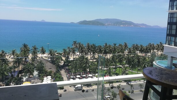 Vista des d'un balcó sobre una platja amb palmeres, una carretera amb vehicles i una serralada de muntanyes a la distància.