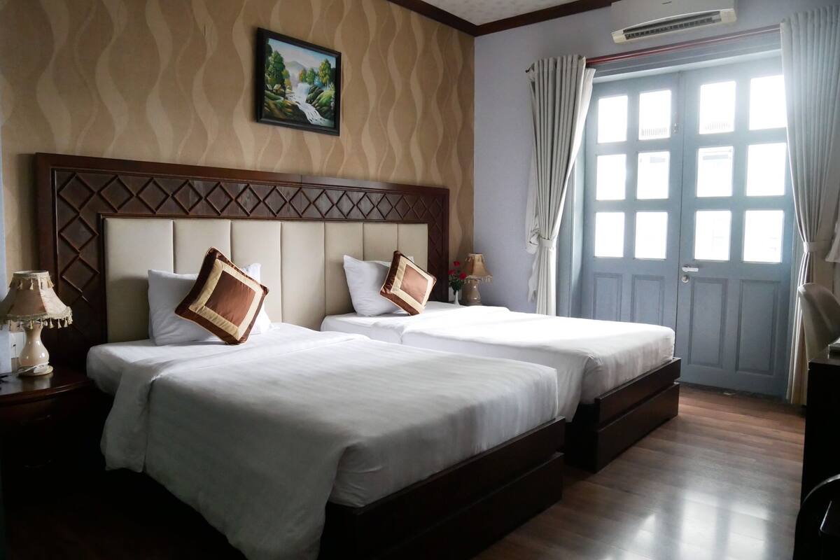 Une chambre d’hôtel avec deux lits, une tête de lit en bois, une table de chevet avec une lampe, un tableau au mur et une fenêtre avec des rideaux.